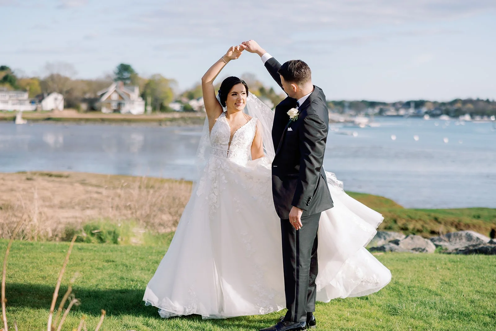 Bride and groom dancing outdoors near water, with the bride in a white wedding gown and the groom in a black suit, overlooking a scenic waterfront with houses and boats.