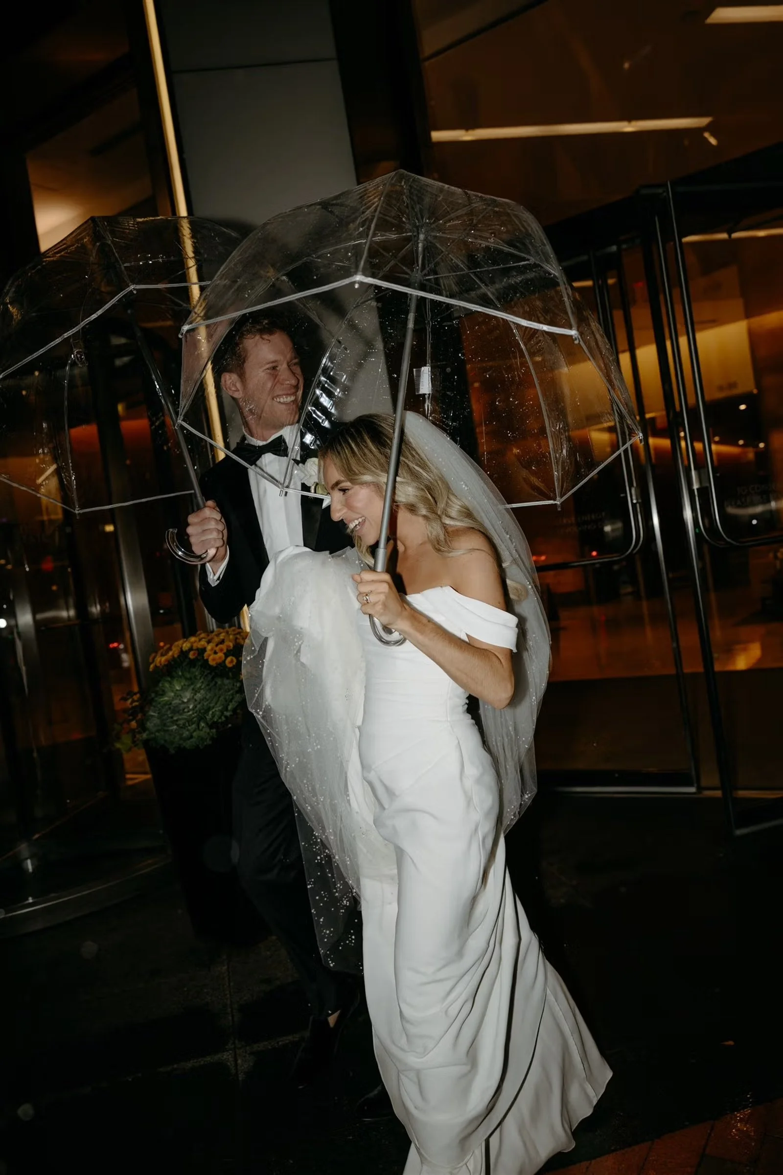 Bride and groom under clear bubble umbrellas on a rainy nigth outside a building with large glass doors.