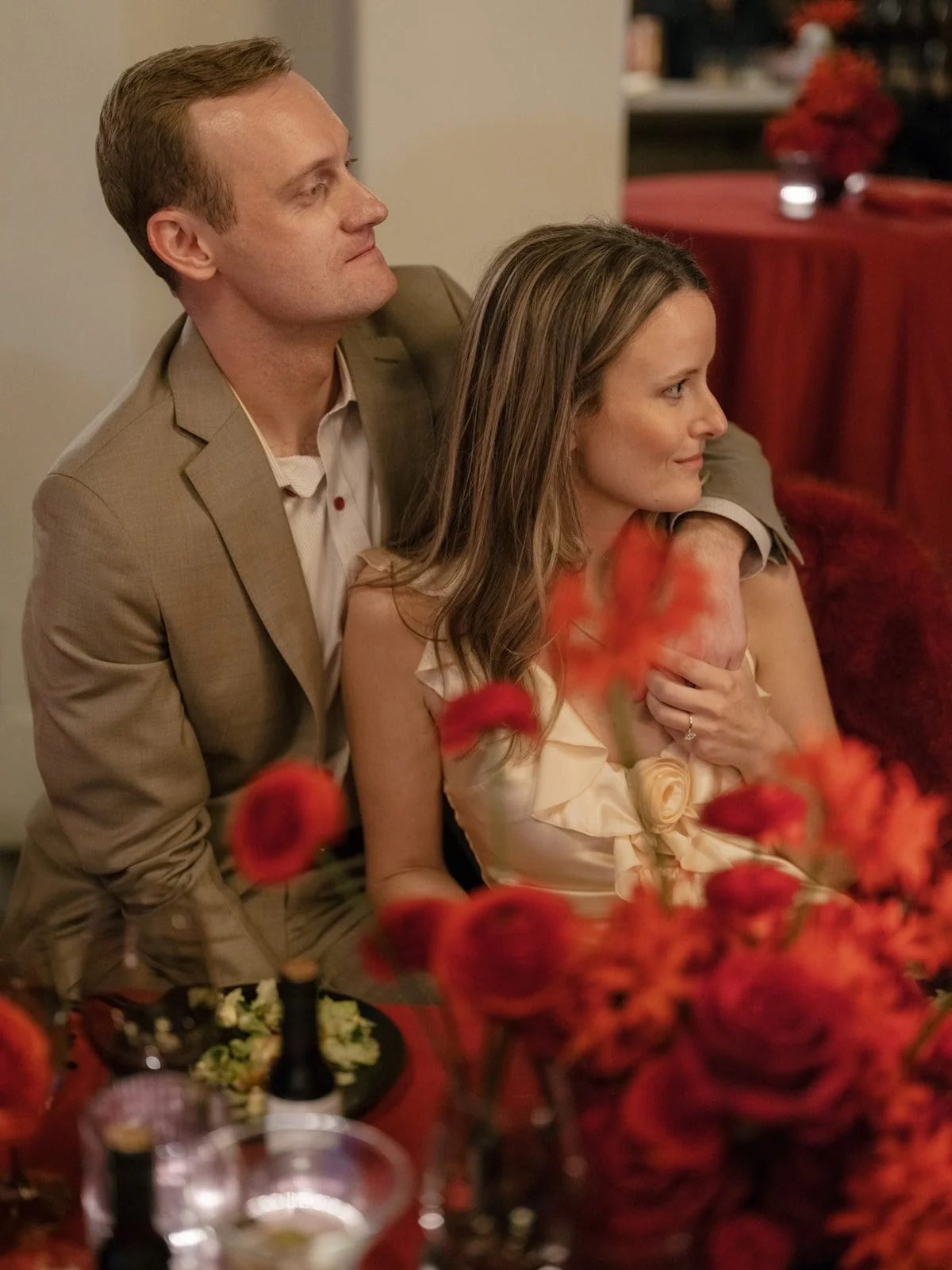 A man in a kaki suit and a woman in a rose gold dress sit togther surrounded by red rosese.