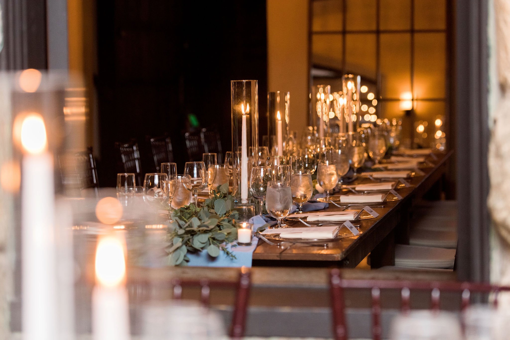 Wedding table set with hurricanes, white tapper candles, wine  glasses, white napkins, silverware, and greenery on a brown farm table, in front of a large paneled window. 