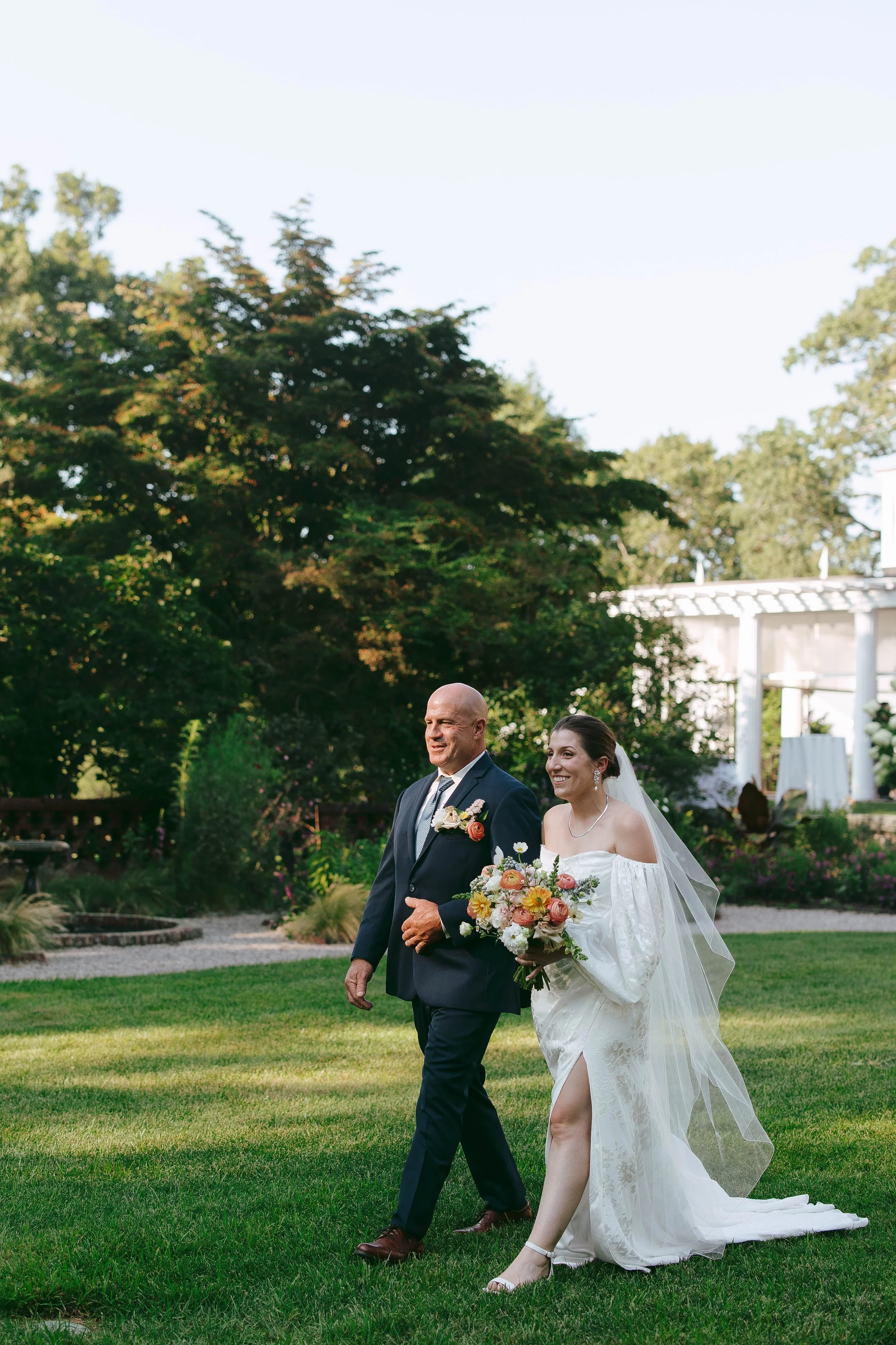Bride walking in a feild past large trees, with a man in a green suit. She is holding a boquest of colorful flowers, with a white building in the background. 