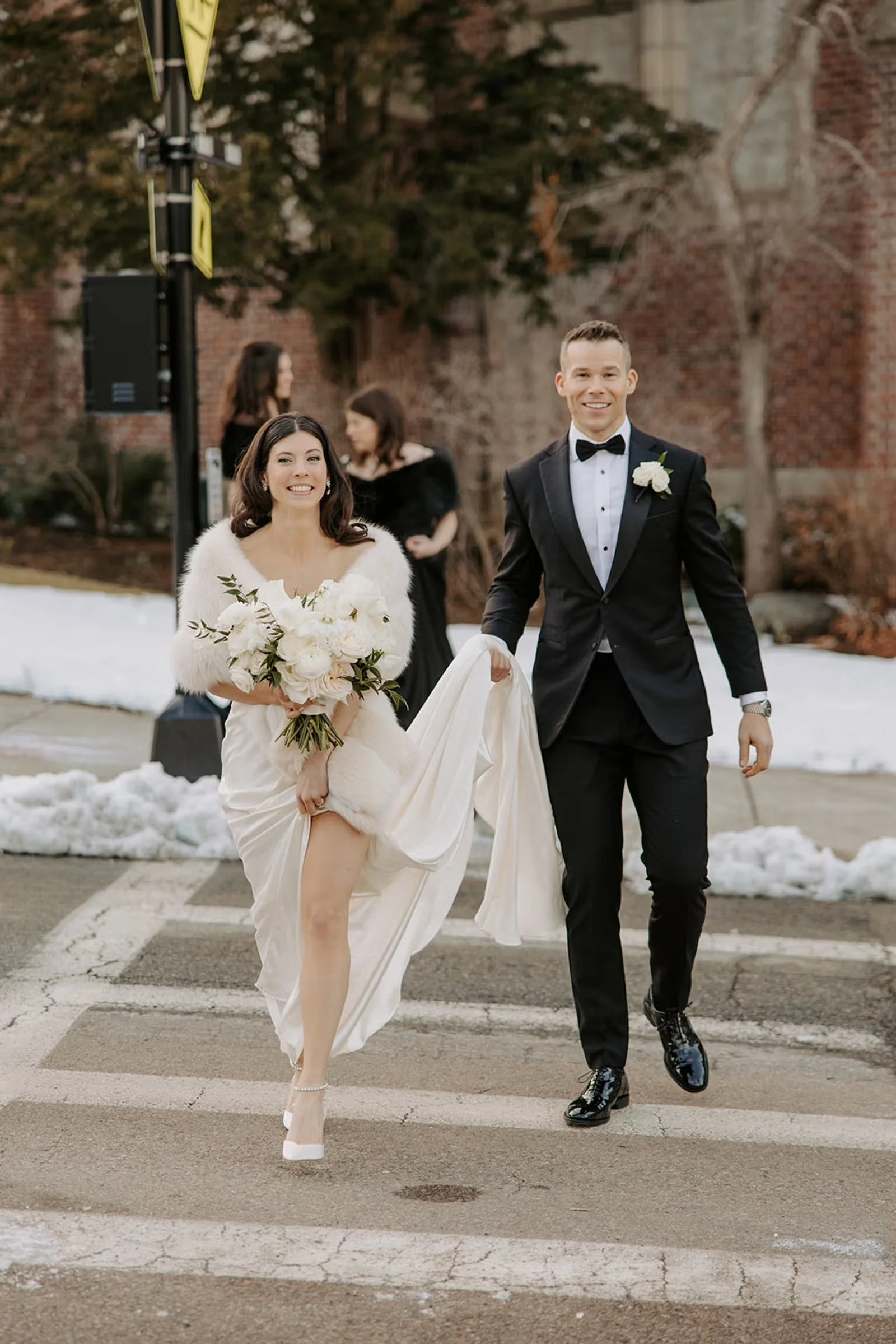 Bride and groom crossing the street. The women is wearing a white dress and winter stole while holding a bouquet of white flowers and the groom is holding up her dress. There is snow on the ground.