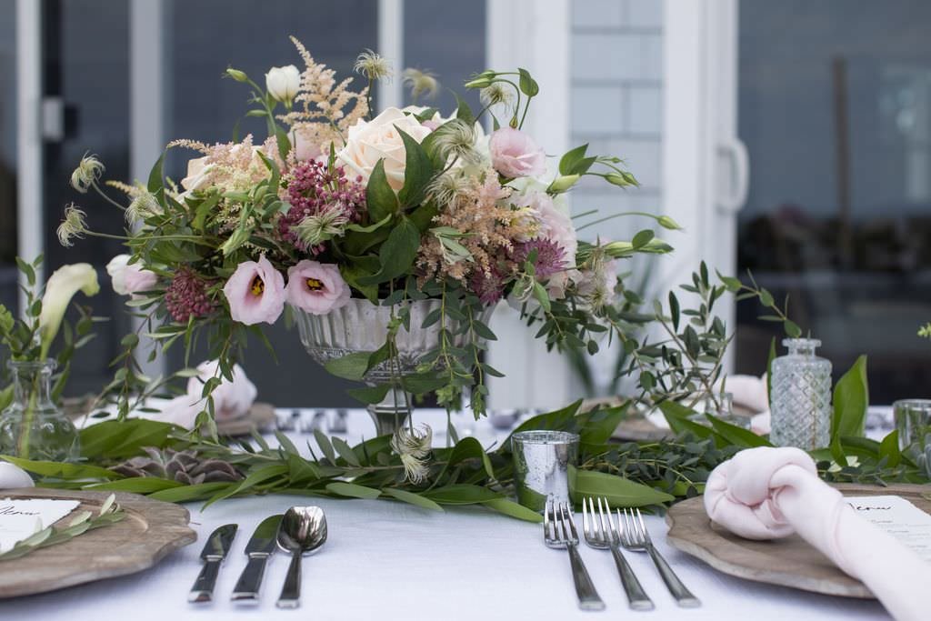 Elegant table setting with a large floral centerpiece featuring pink and white flowers, greenery, and feathers. The table has a white tablecloth, silverware, and wooden chargers, with napkins and glassware in a sophisticated dining arrangement.