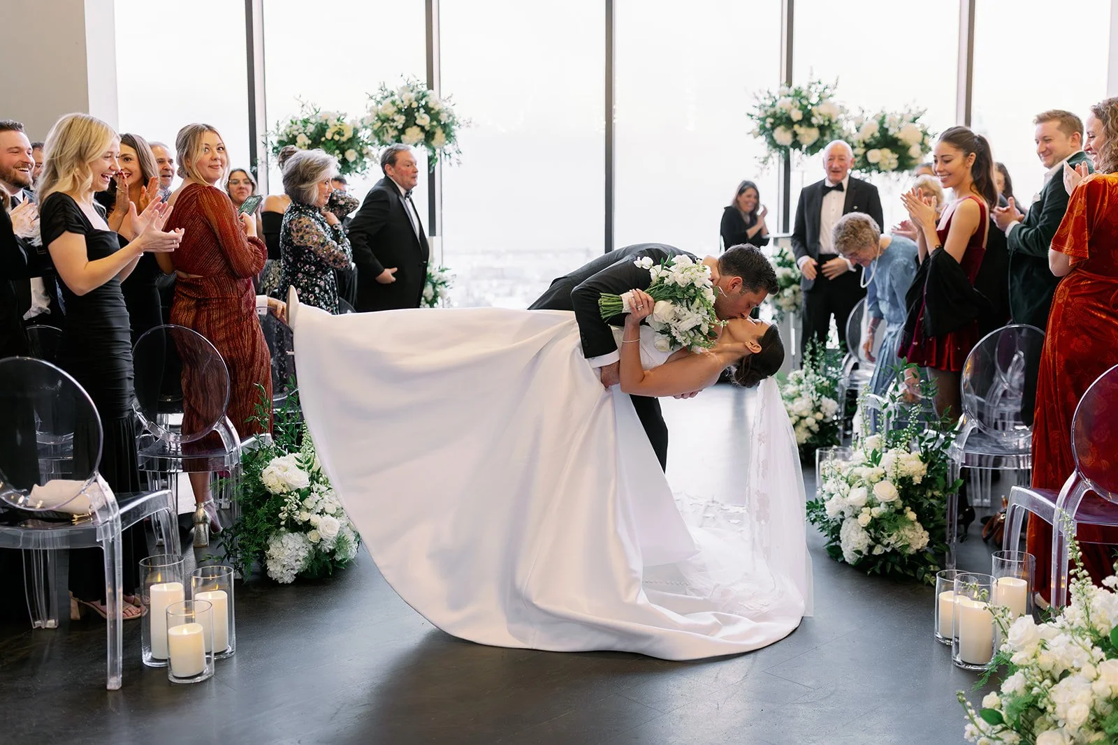 A groom in a black suit dips a bride in a white dress into a kiss at the end of a wedding isle. They are surrounded by guests and green and white florals as well as white candles.