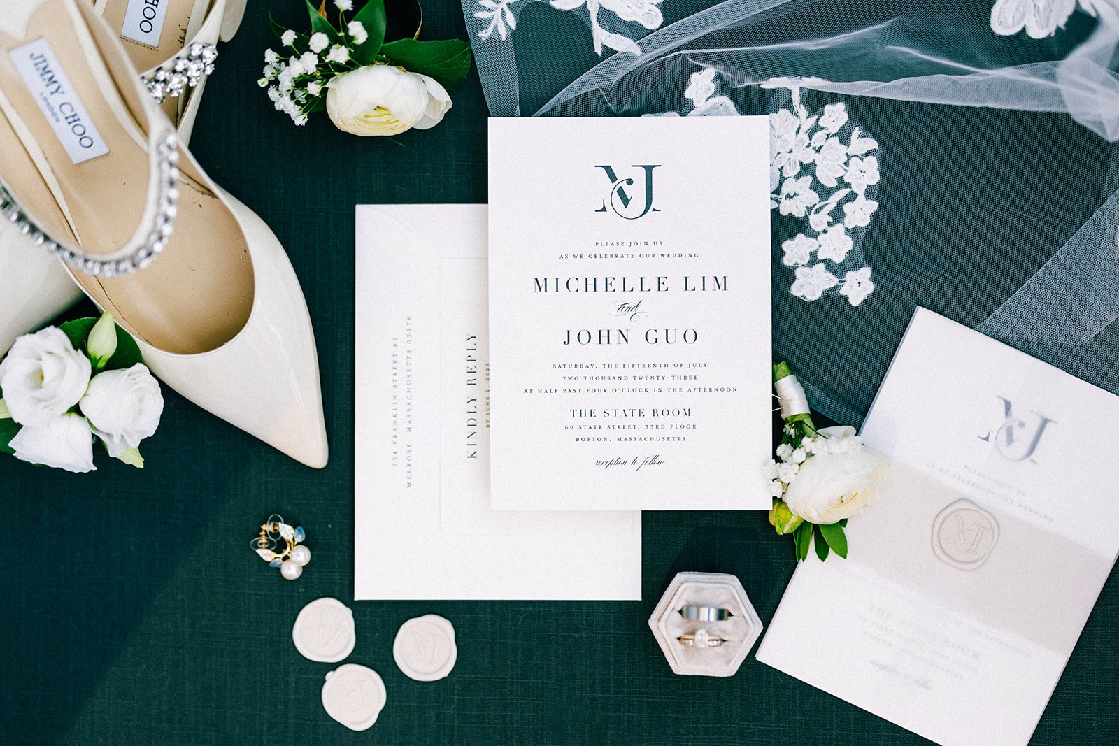 Wedding invitation suite with floral accents, white shoes, jewelry, and wedding accessories on a dark surface.