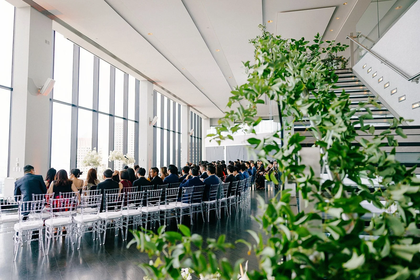 Wedding ceremony framed by casading greenery and flowers, with guests seated in acrylic chairs beside floor-to-ceiling windows.