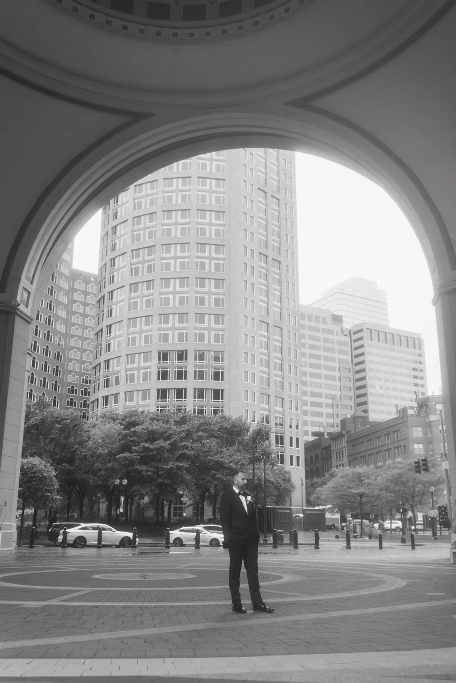 Man in a suit stannding under an arch in city park with a large sky scrapper in the background.