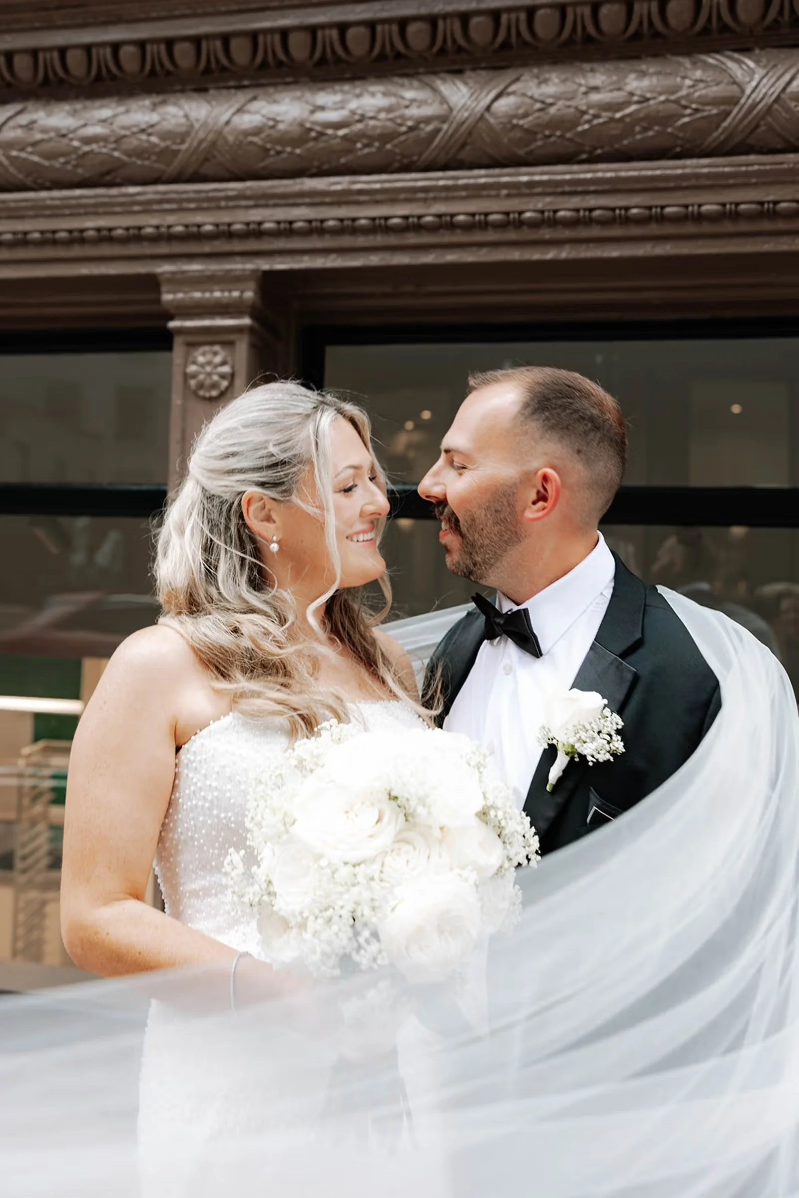 A bride with a long veil holds a colorful bouquet of flowers and holds the grooms suit.