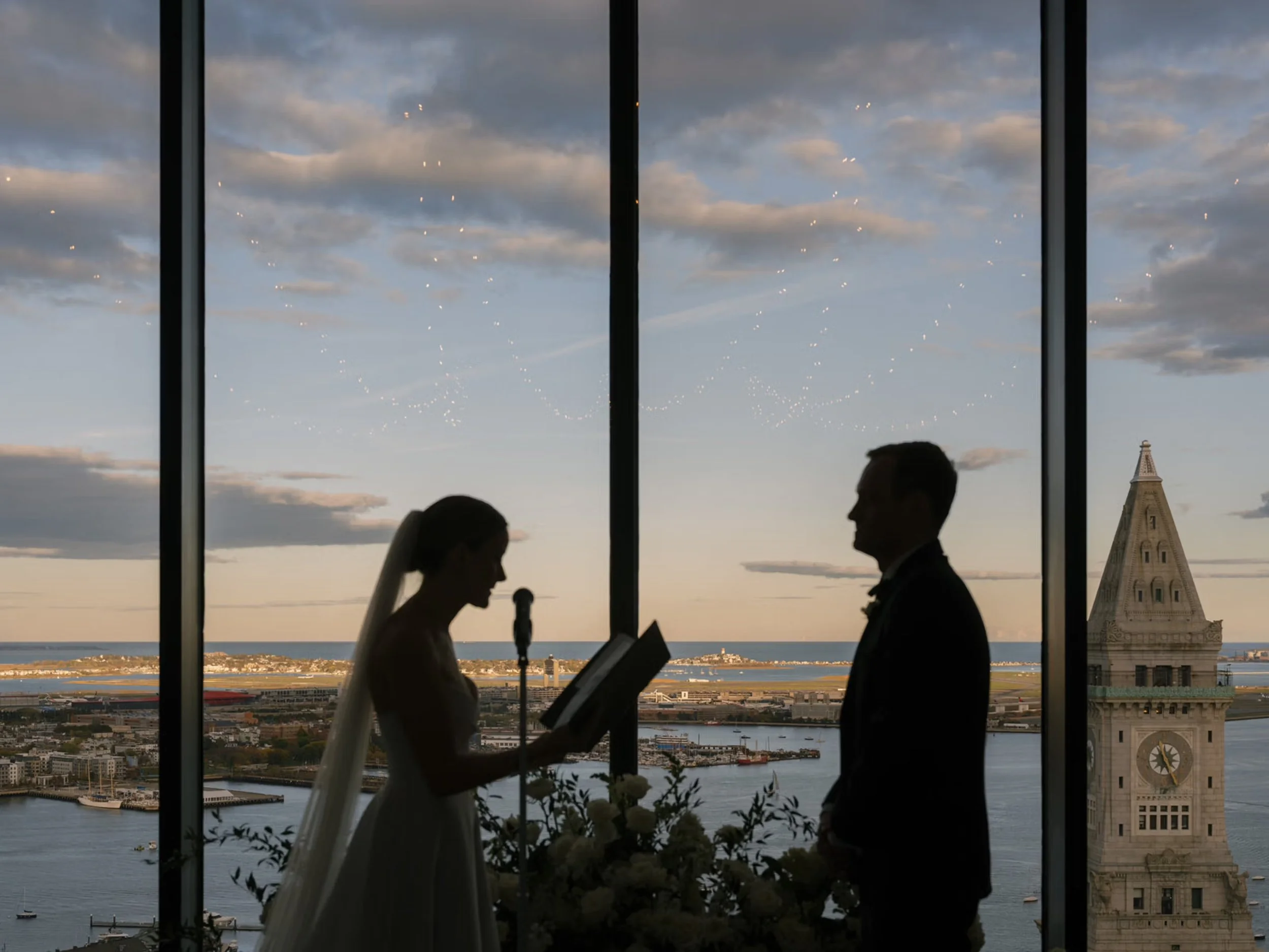 Bride and groom backlit in front of a large window and a floral arrangment with a clock tower in the background at dusk with clouds scattering the sky.