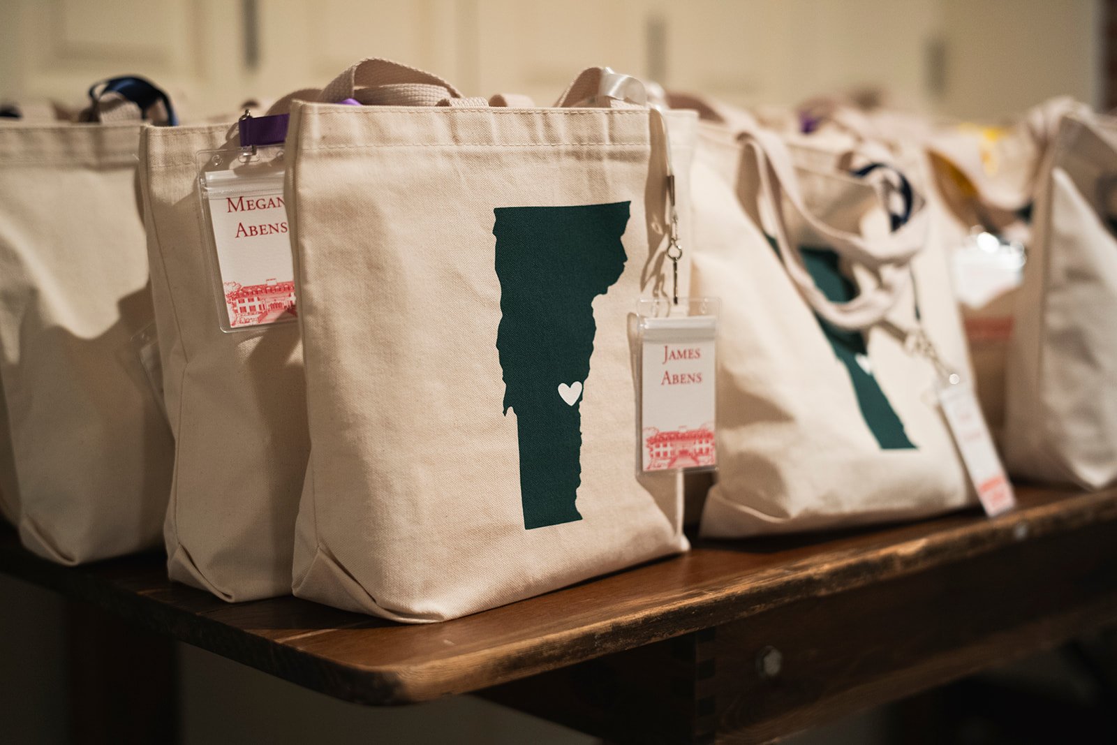 A wooden table is lined with kaki canvas totes with a green print of Vermont.