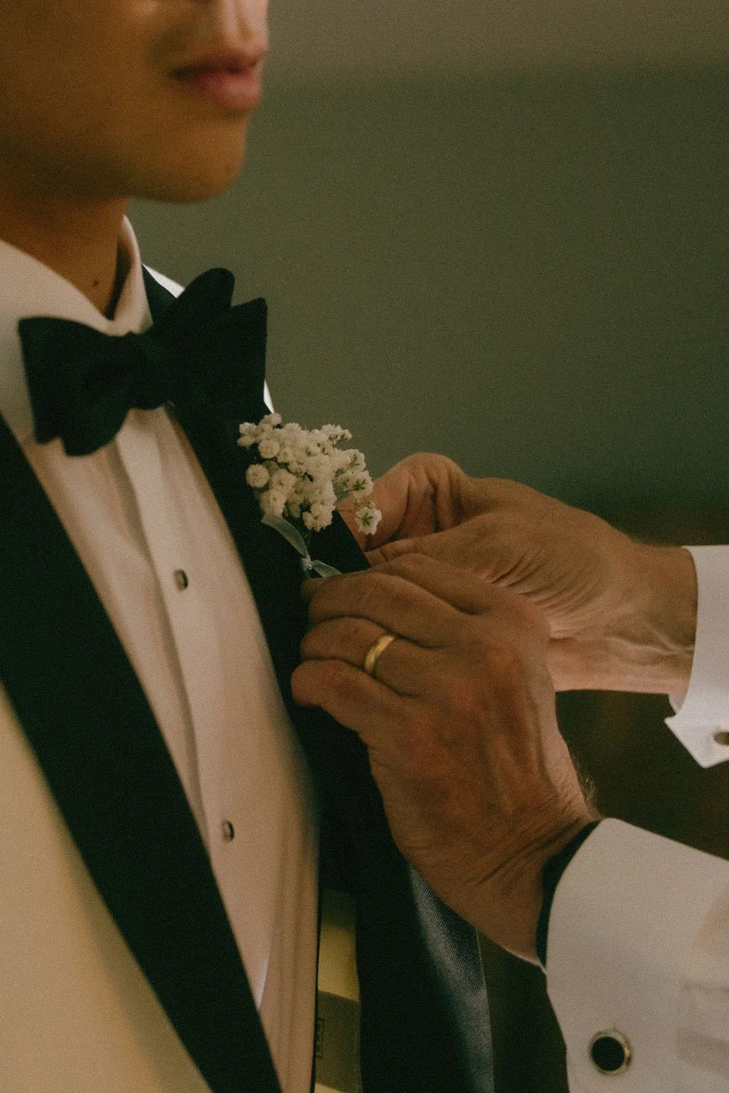 Man with a gold wedding band pinning a white boutonnière on a man in a white and black tux with a black bowtie 