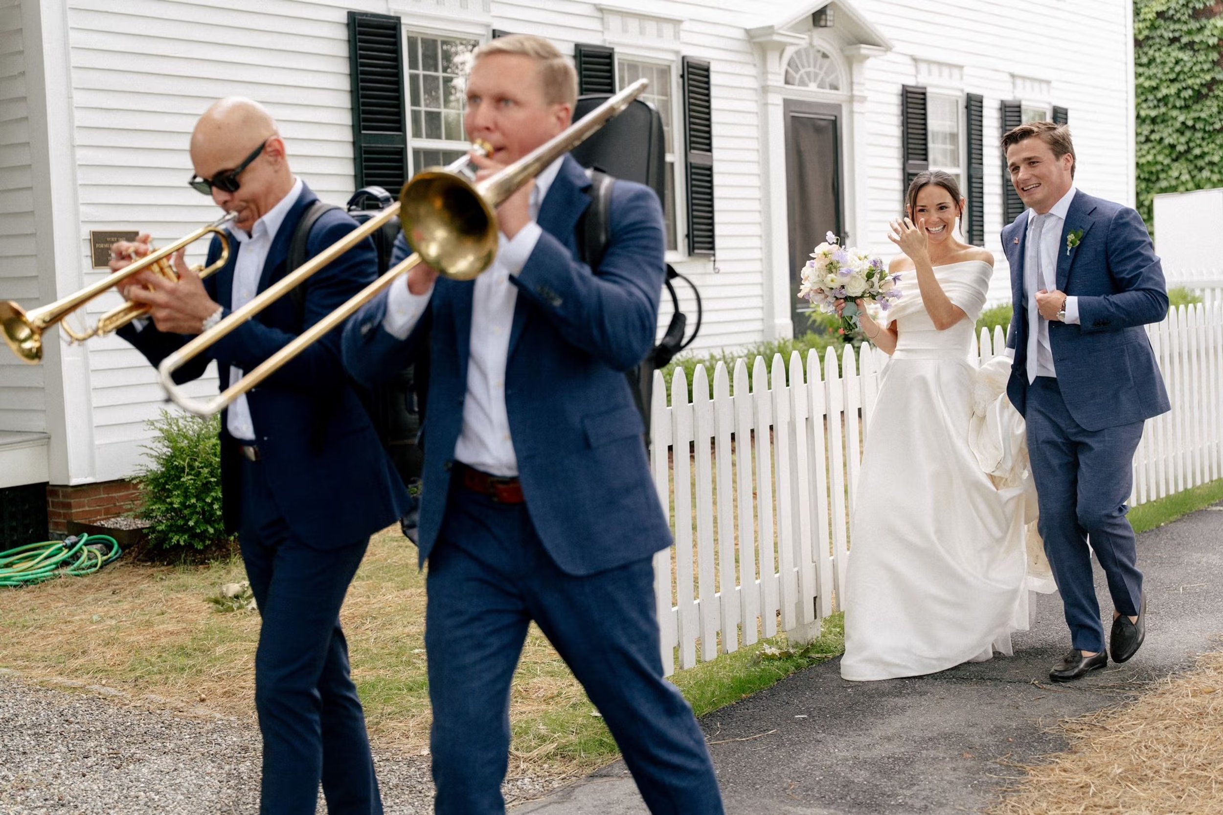 Bride and groom walking down the sidewalk. Passing by a historic white house, and following men in blue suits playing trumpits. 