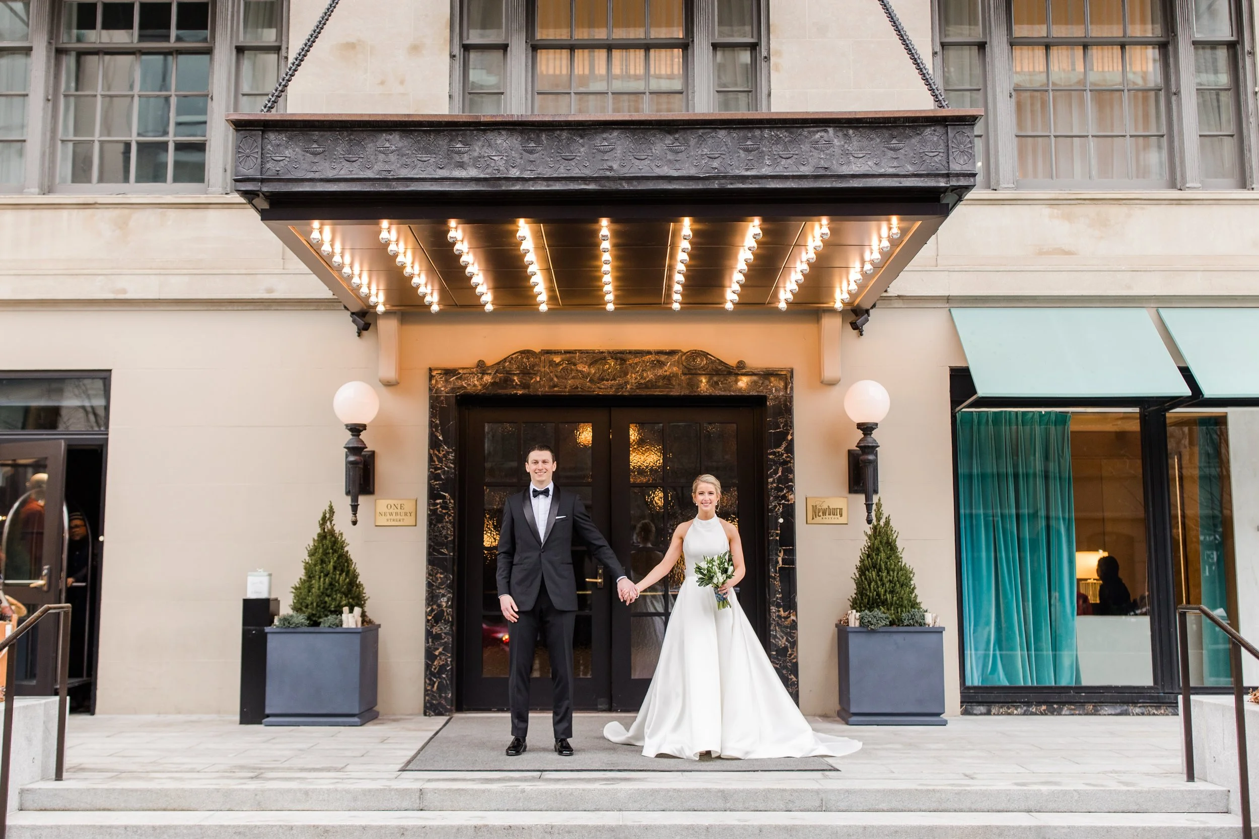 A bride in a white dress holding a white boquet holds hands with the grrom in a balck suit and bowtie. The bride and groom are standing infront od the main entrence to a downntown hotel with double doors, a large window, and canopy lights.