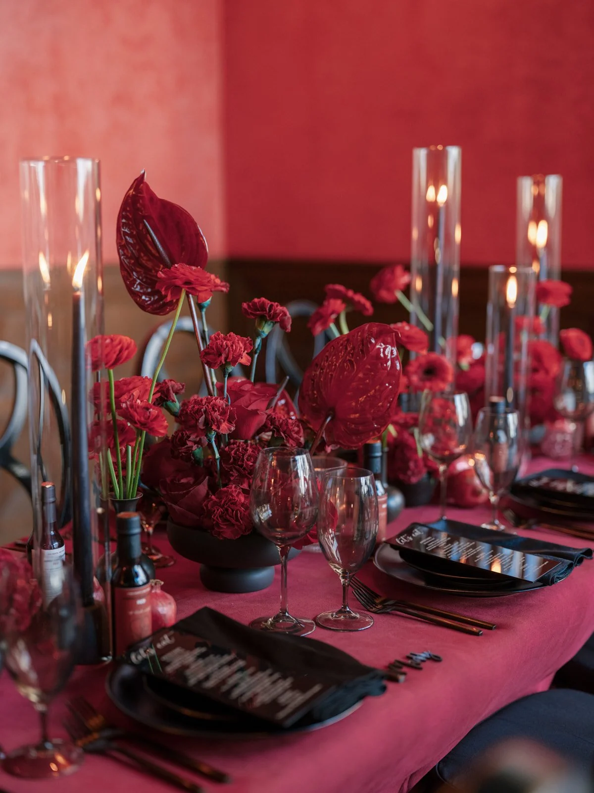A table set for a celebration with a maroon tablecloth, floral centerpieces that showcase deep red Calla lilies, brick red tapper candles, and place settings with black napkins.