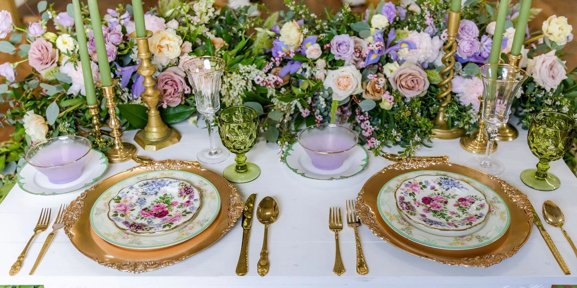 Elegant table setting with floral plates, gold cutlery, green and purple glassware, golden candlesticks, and a large floral centerpiece of pink, purple, and white flowers.