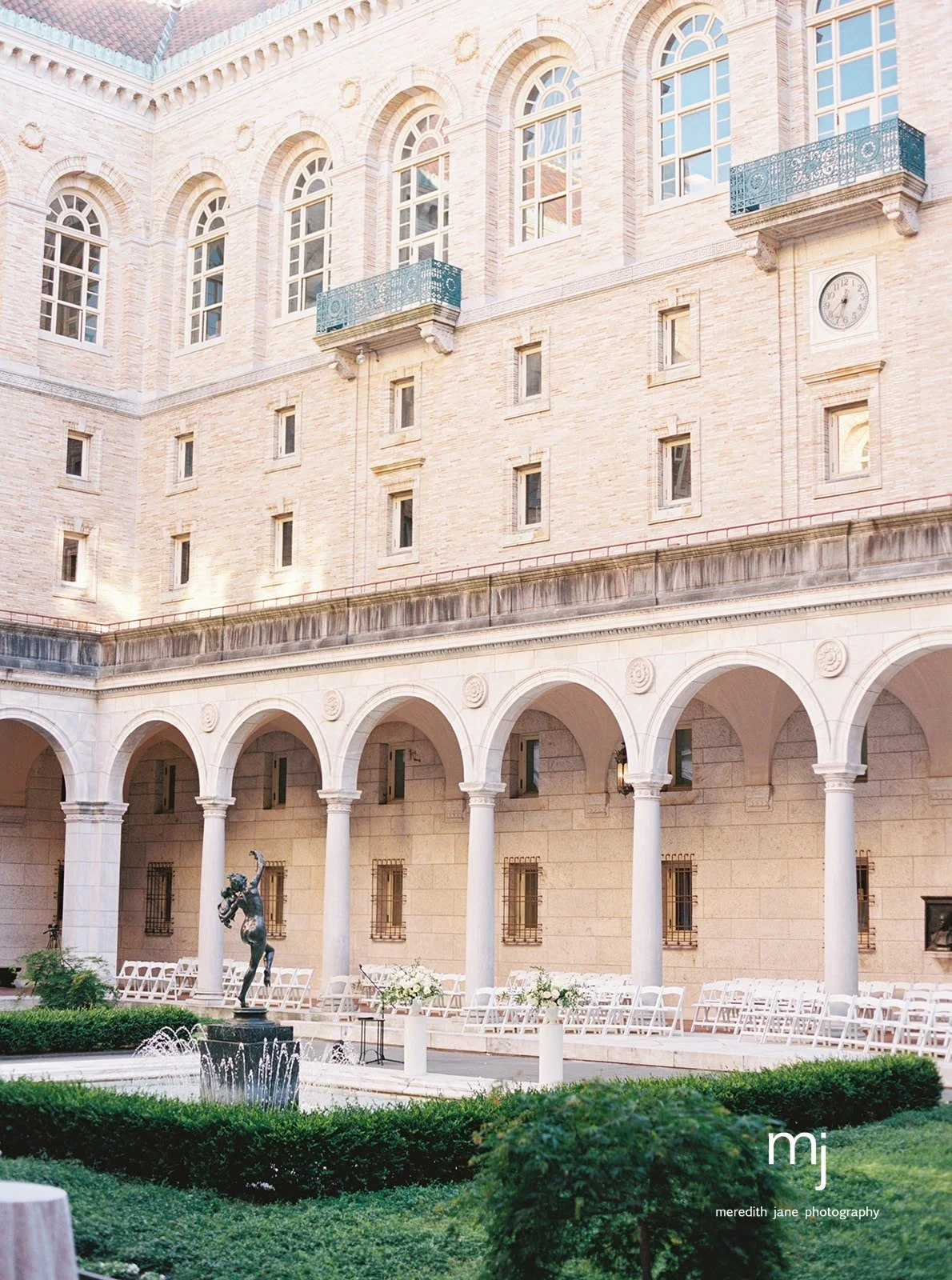 The image shows the exterior of a large, elegant building with light-colored brick walls and multiple arched windows. There are two small balconies with blue railings on the upper part of the building. On the ground level, there is a covered patio area with white chairs, a small fountain with a statue in the center, and some white floral arrangements. The setting appears to be prepared for an outdoor event or gathering, surrounded by well-maintained greenery.