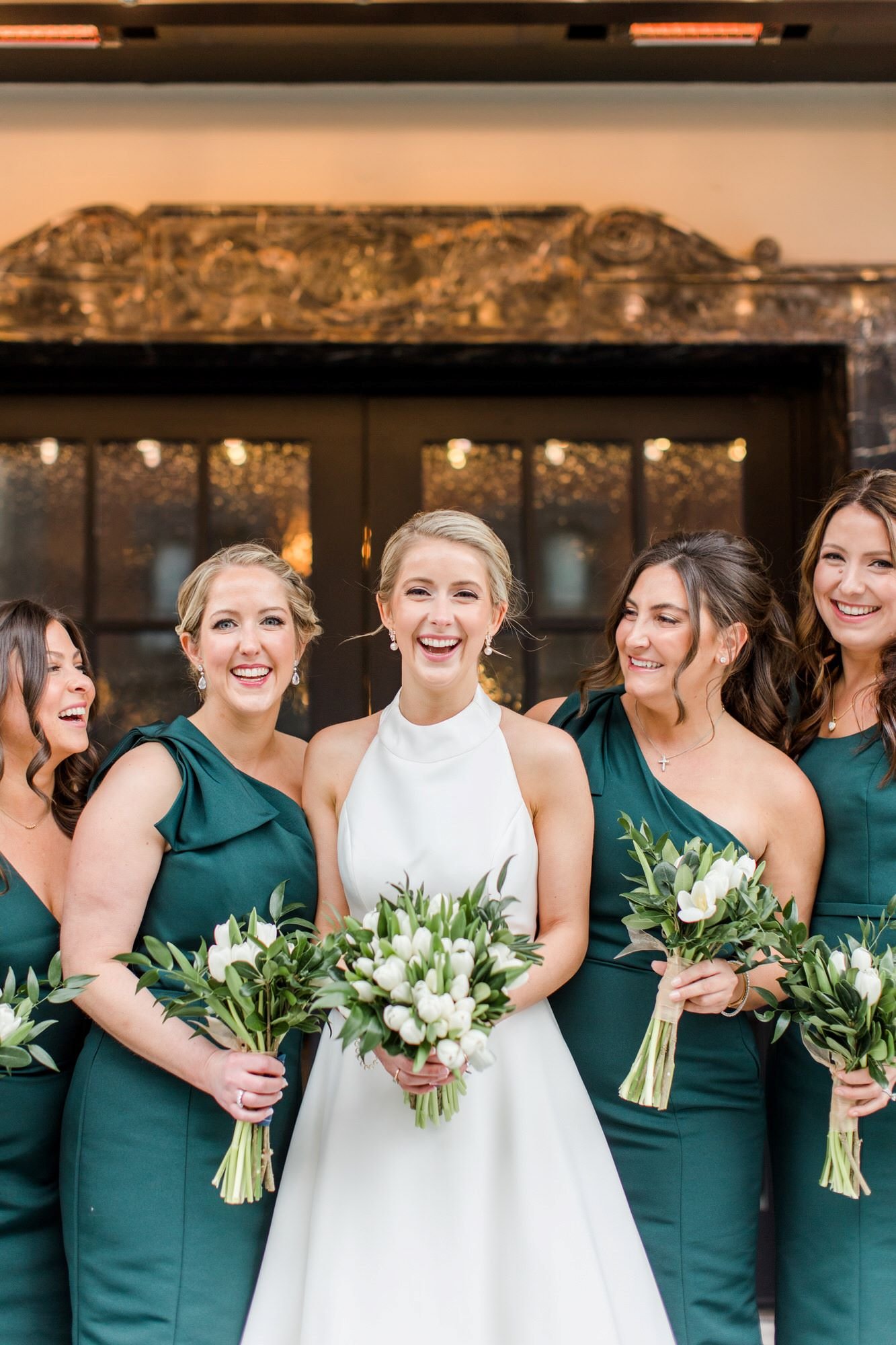 A bride in a white dress standing with her bridesmaids in dark green dresses, holding bouquets, smiling at a wedding celebration.