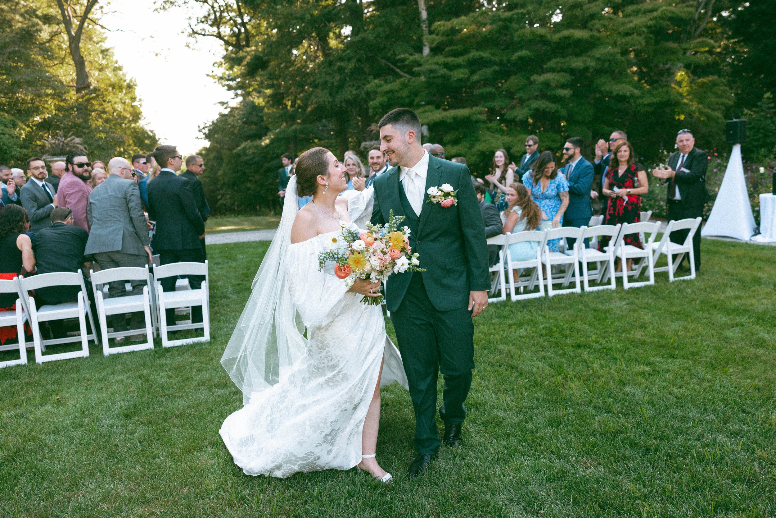 Bride and groom walking together at an outdoor wedding ceremony, with guests seated and clapping in the background, surrounded by green trees on a sunny day.