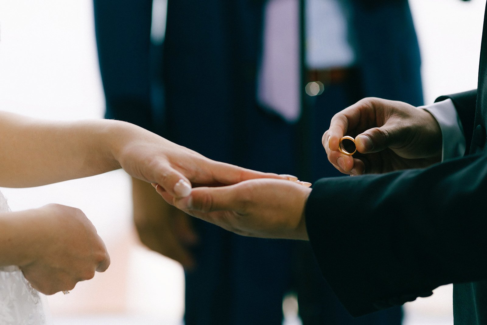 A groom in a black suit slips a gold wedding band on the brides hand.
