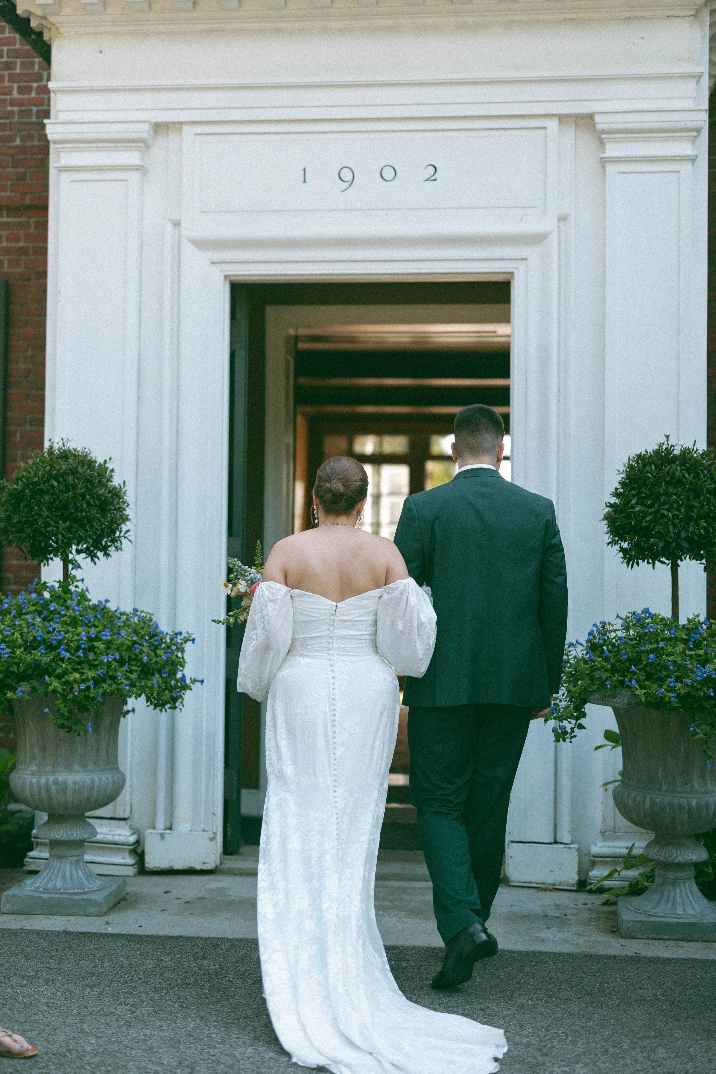 Women in a long whote dress and groom in a green suit entering a white building next to potted topiary plants.