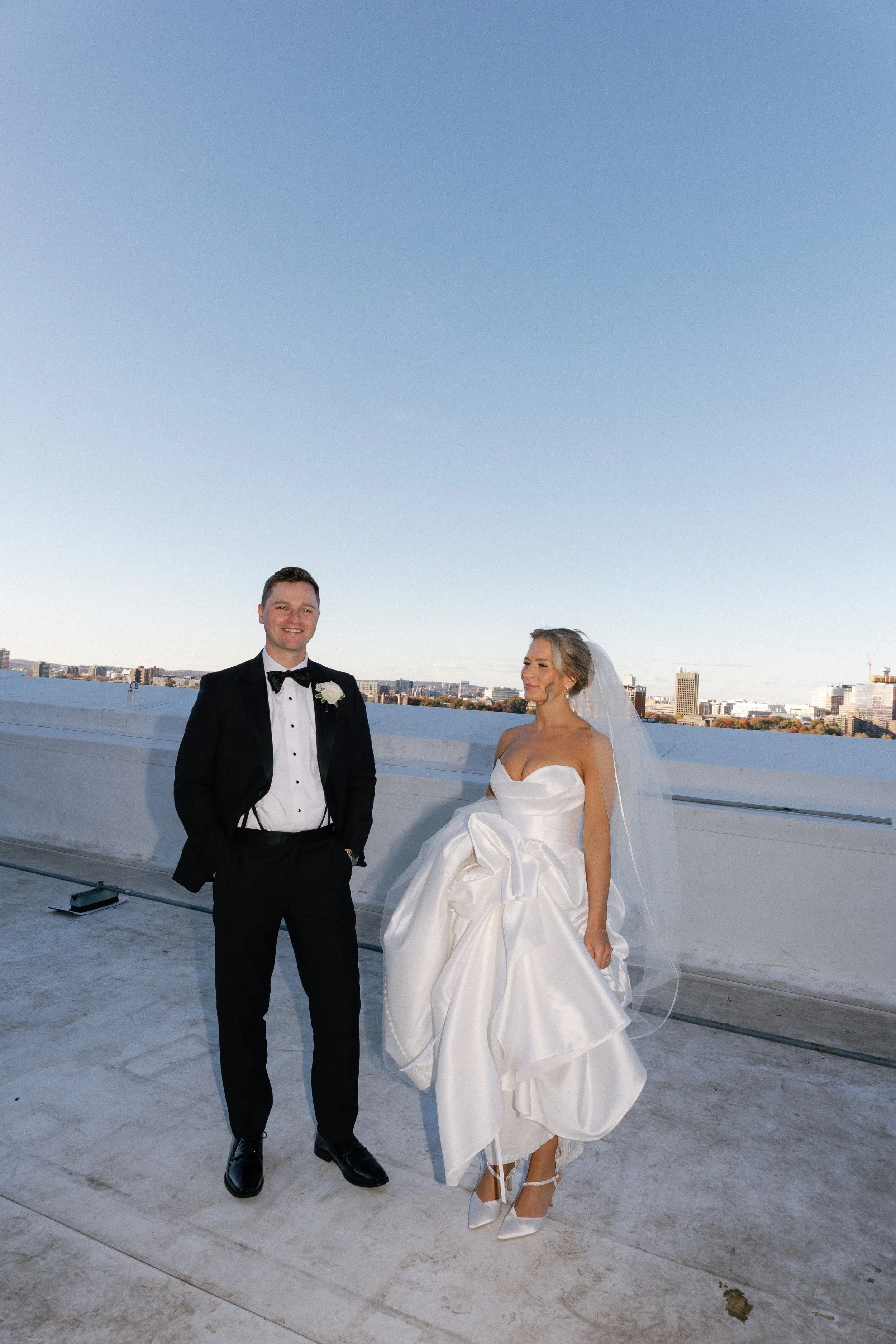 a bride in a white dress and a groom in a black tux stand infront of a city sky line featuring the ocean.