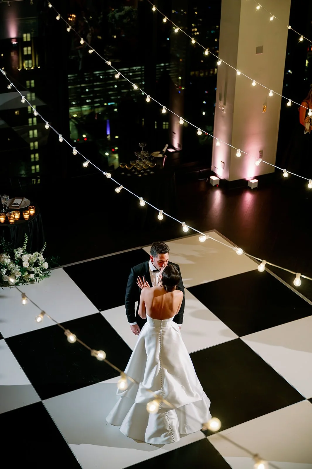A bride in a white dress kisses a groom in a black tux while dancing. They are dancing on a black and white checked dance floor under Edison bulb string lights.