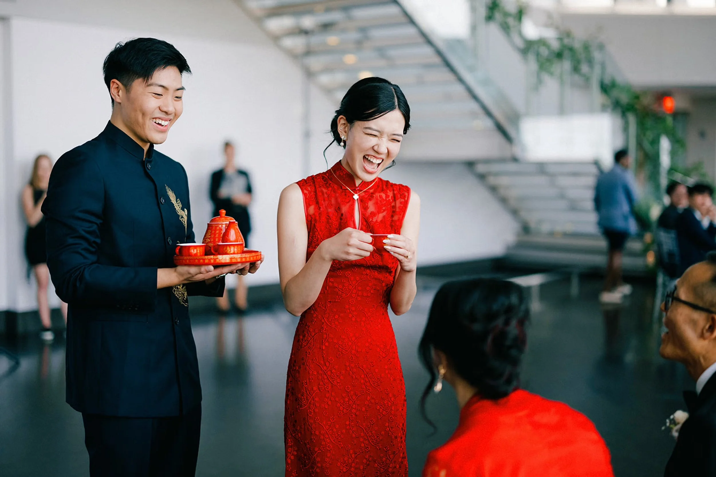 Couple participating in a traditional Asian tea ceremony, with the bride in a red dress, with the groom carrying a red ceremonial tea set.