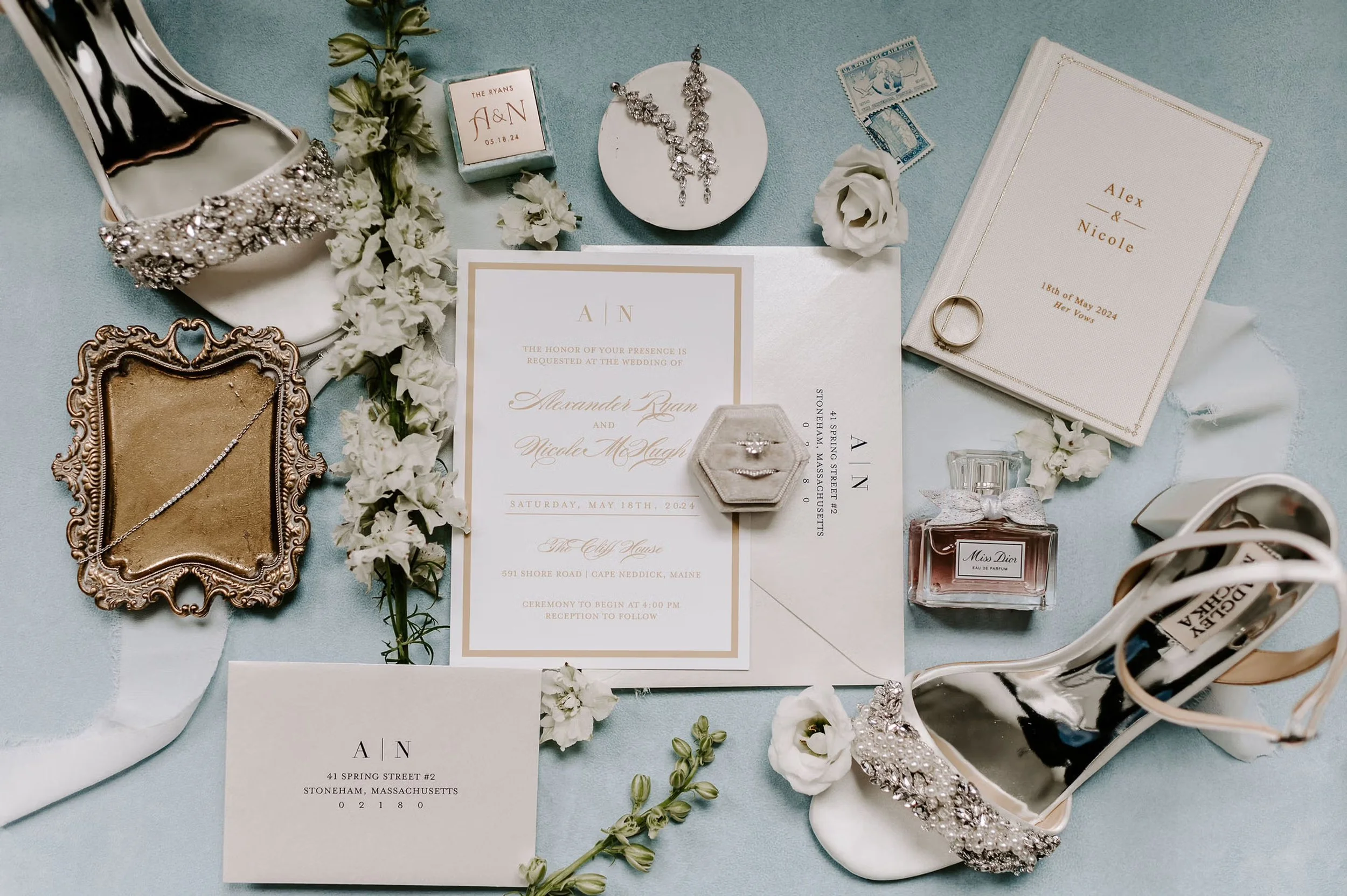 Wedding invitation suite with floral accents, white shoes, jewelry, and wedding accessories on a light blue backdrop.
