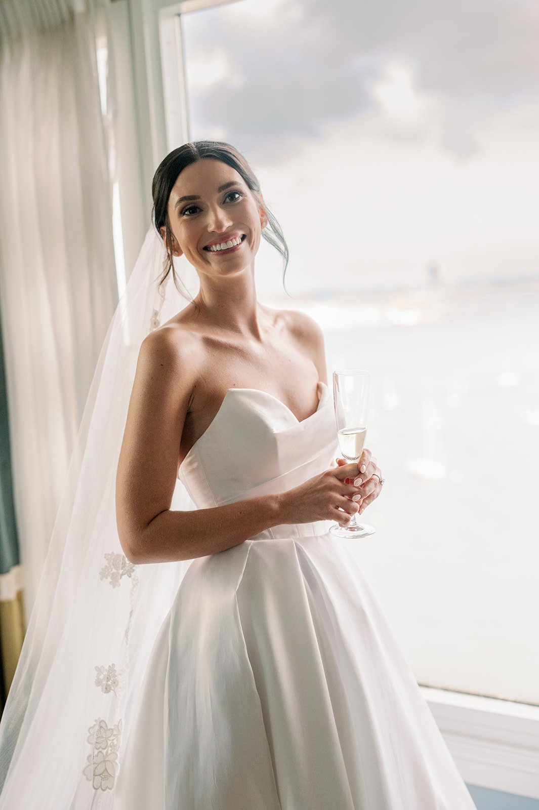 Bride in white dress and veil with lace details smiling in front of a large window.