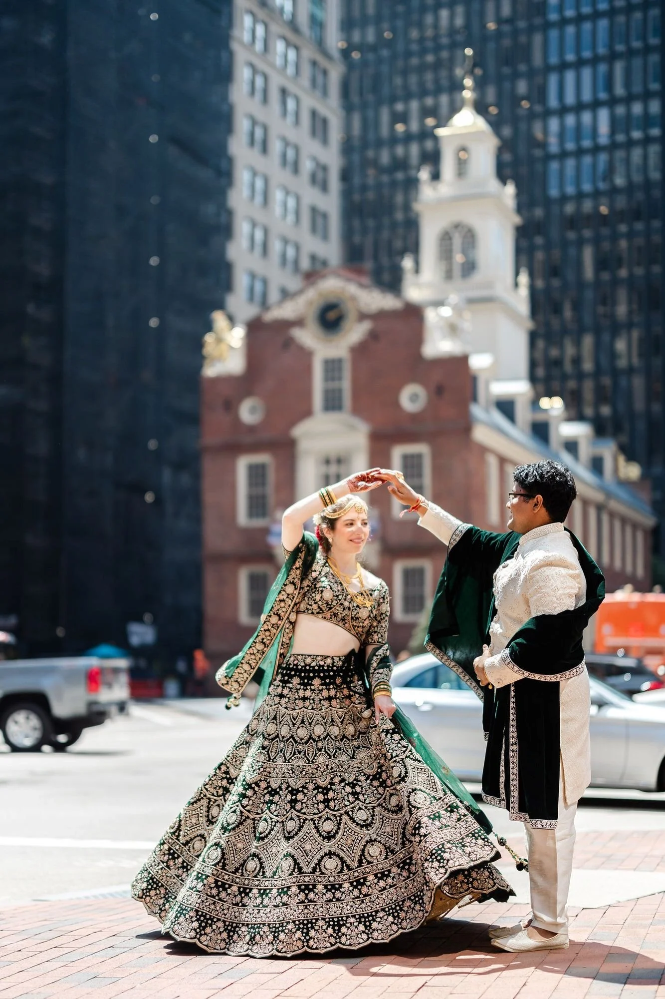 A couple dressed in traditional Indian wedding attire dancing outdoors in an urban setting with skyscrapers and a church building in the background.