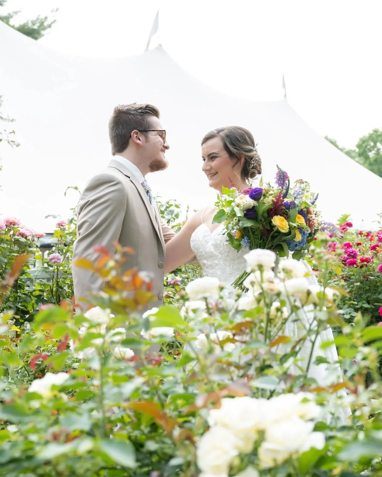A groom in a tan suit and a bride in a white dress holding a boquet of colorful flowers, standing in a feild of wildflowers.