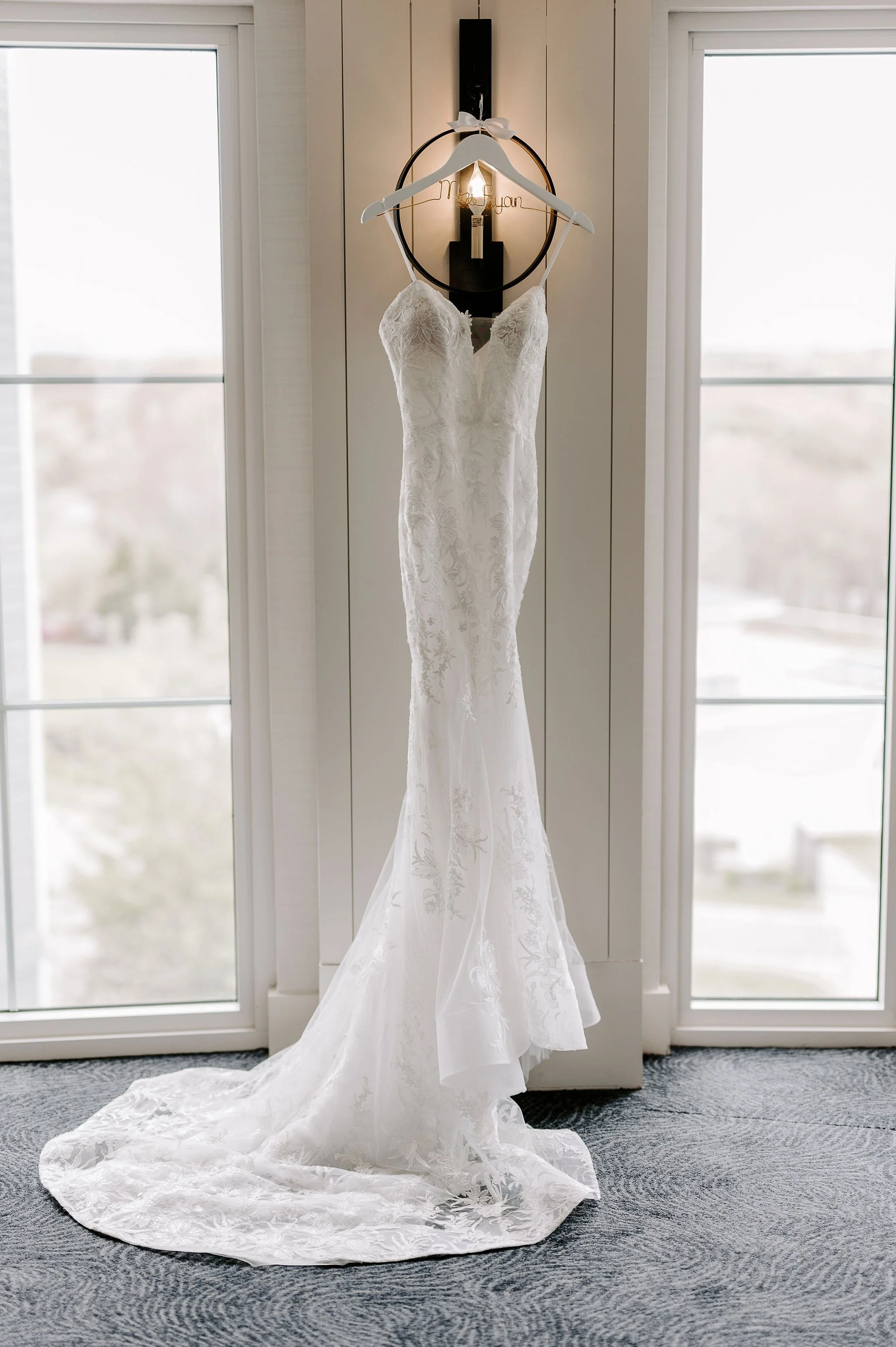 White wedding dress with a long train hangs on display in front of a white wall and large windows.