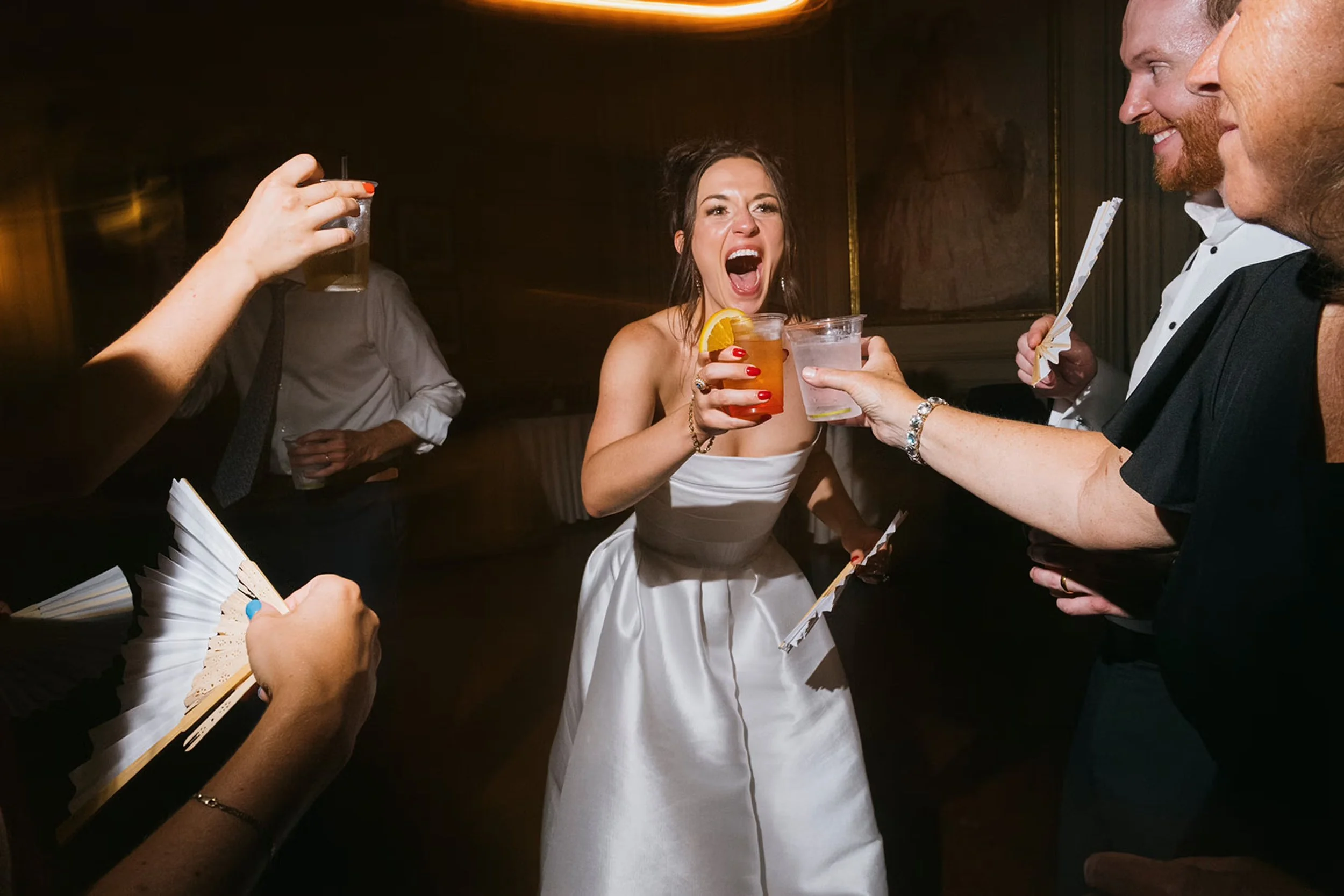 Women in a white dress holding a red cocktail while guests cheers around her at a wedding reception.