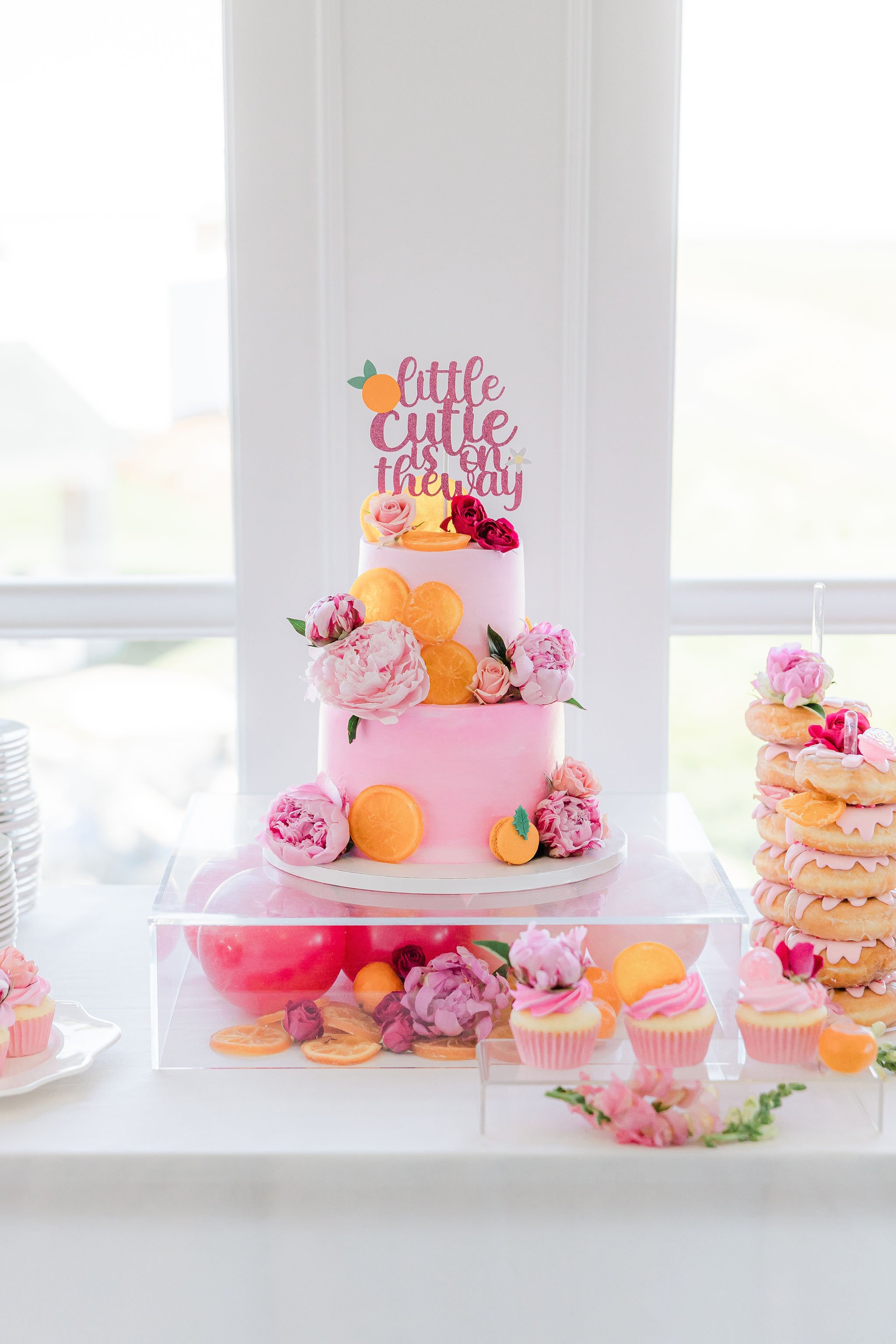 A pink two tiered cake is decorated with pink peonies and orange slices, the cake sits on an acrylic cake stand filled with hot pink balloons. To the side of the cake is a stack of pink frosted donuts.