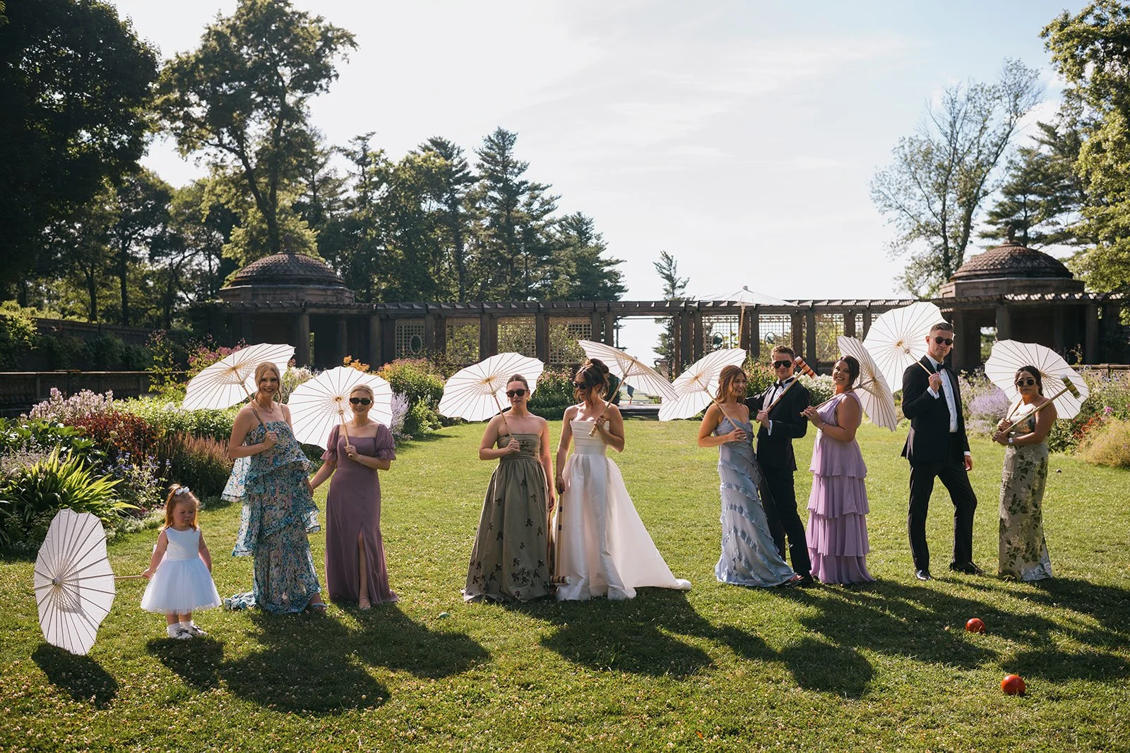 Wedding party in spring dresses and suits standing in a grassy feild with paper parasols and stone structures in the background.