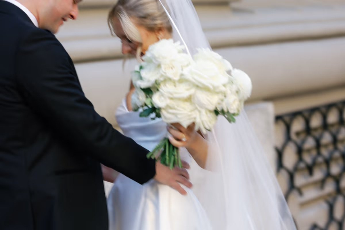 Groom holding a bride in a white dress and veil while she holds a bouquet of white roses in front of a stone building. 
