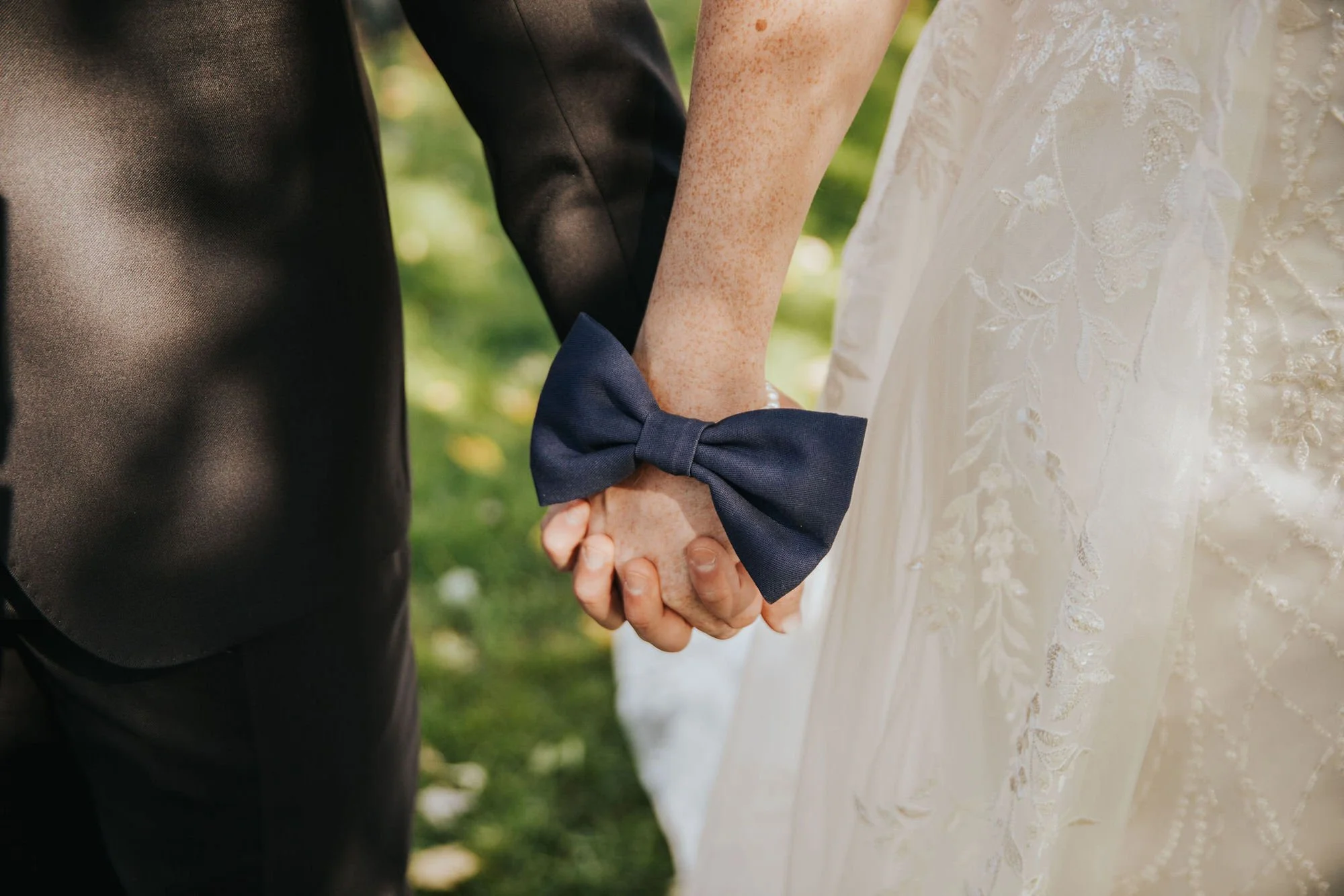 Close-up of a bride and groom holding hands, with the bride wearing a wedding dress with embroidery and a navy bow bracelet on her wrist.