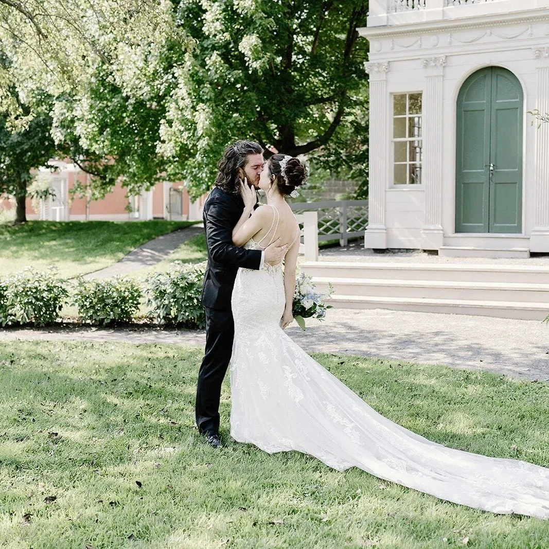 Woman in a white dress kisses a man in a black suit. They are kissing front of a large green tree with a white building in the background.