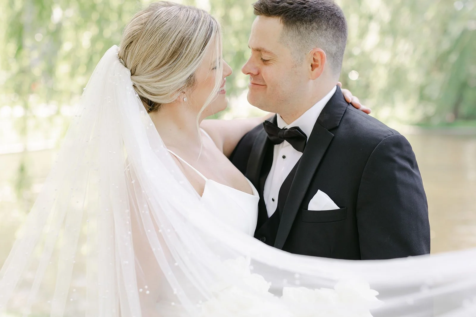 Bride and groom sharing a close moment outdoors, bride in a white dress with veil, groom in a black tuxedo with bow tie.