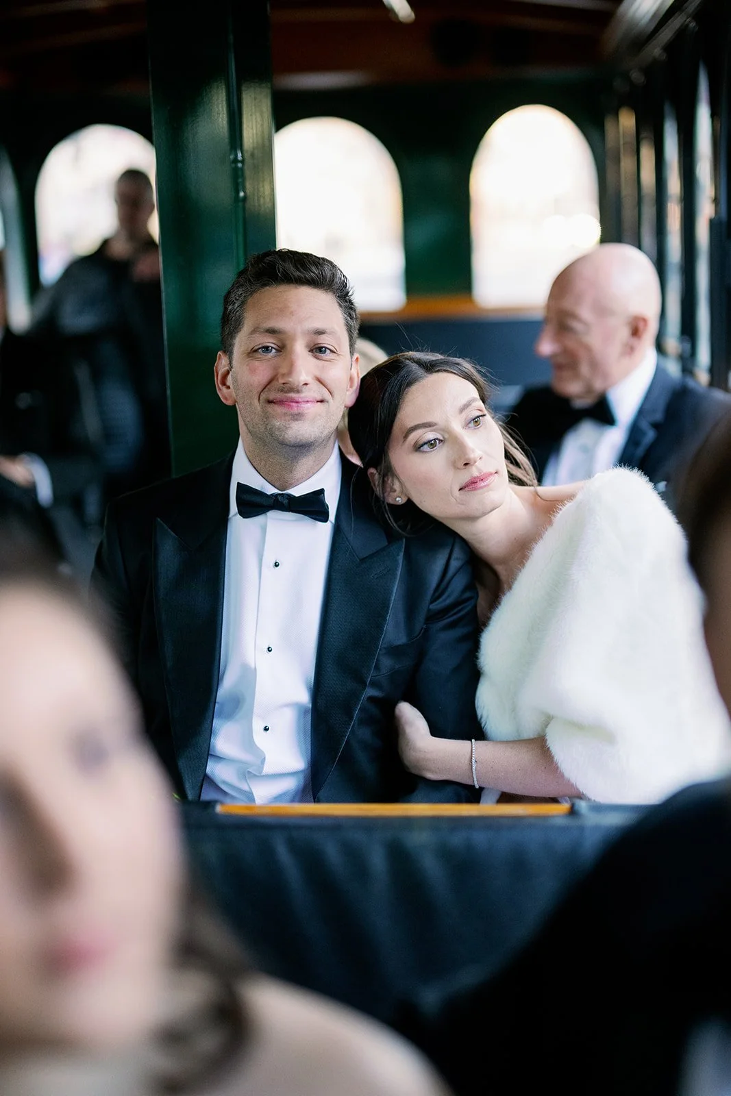 Bride wearing a fur stole leans her head on the groom in a black bowtie while they ride in a trolly surrounded by other members of the bridal party. 