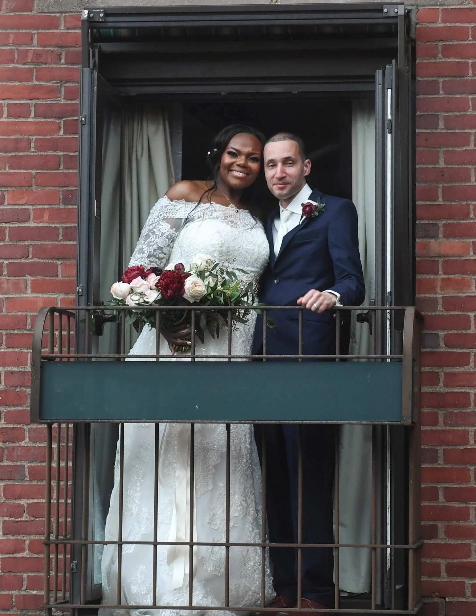 A bride and groom standing on a small balcony, looking at the camera. The bride wears a white lace wedding dress and holds a bouquet of red, pink, and white flowers. The groom wears a navy blue suit with a white shirt and a boutonnière. They are smil