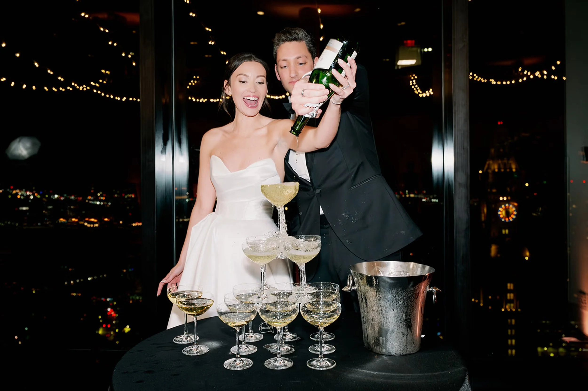 A bride and groom pour a bottle of champagne into the top glass of a champagne tower. They are standing in front of three windows at night time with lights reflecting in the windows and a clocktower in the background.