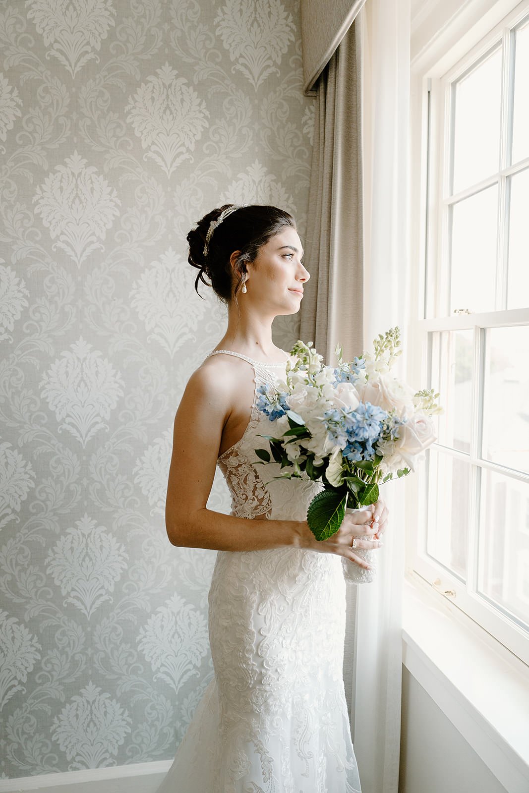 Bride standing by window, holding a bouquet of white and blue flowers, wearing a lace wedding gown, with an updo hairstyle and pearl earrings.