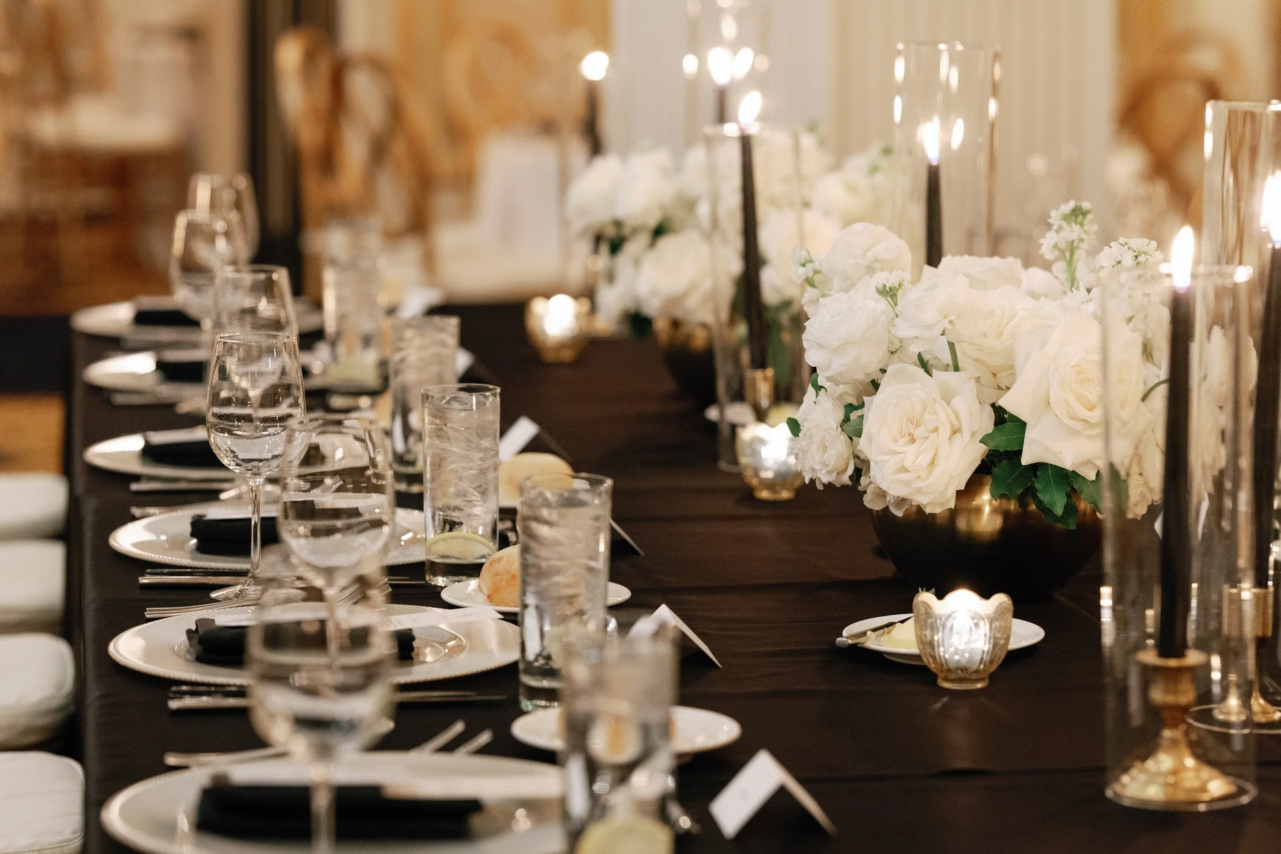 A black banquet table is set with white plates, black napkins, glassware, black tapper candels in hurricans, white floral center peices, and tea lights.