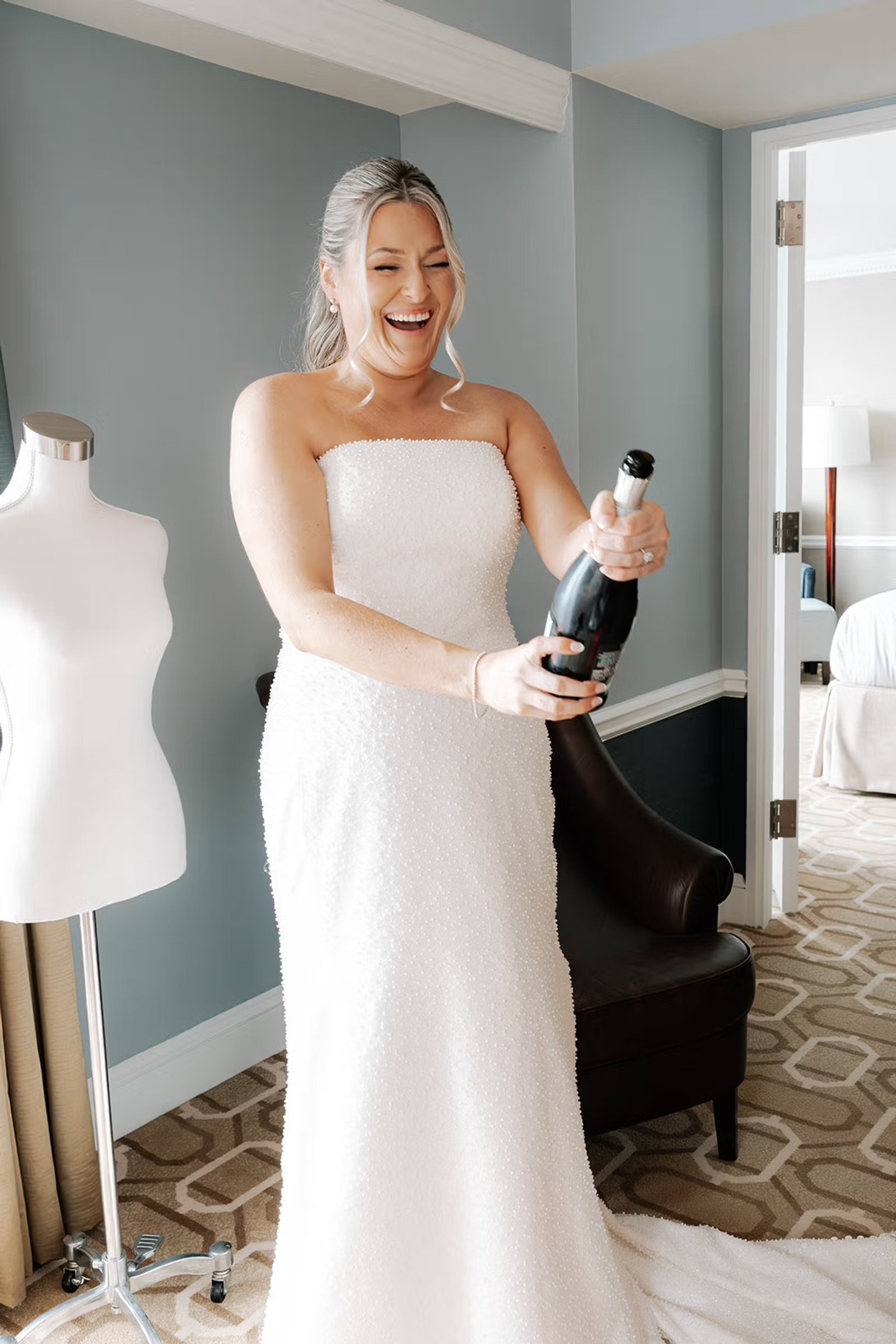 Women in a white dress popping a bottle of champagne in a hotel room, with a white dress form in the background. 