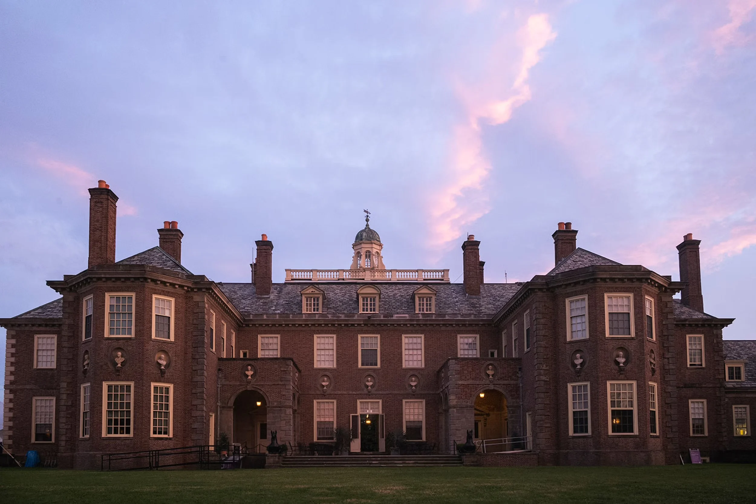 Exterior of a large, elegant, historic building with brick walls and lots of windows. There is a light purple sunset sky in the background.