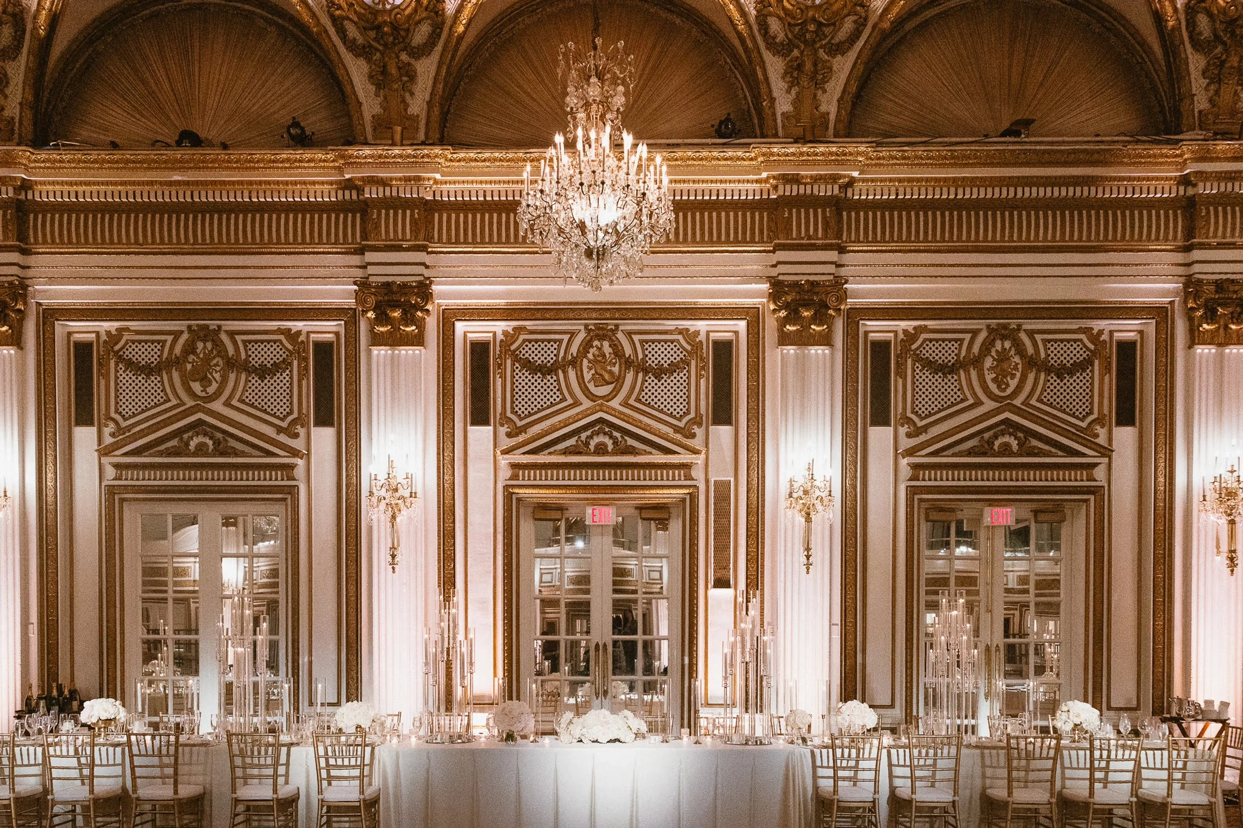 Tables set for a formal event with floral centerpieces, white tablecloth, candles, glasses, and silverware, near large window.