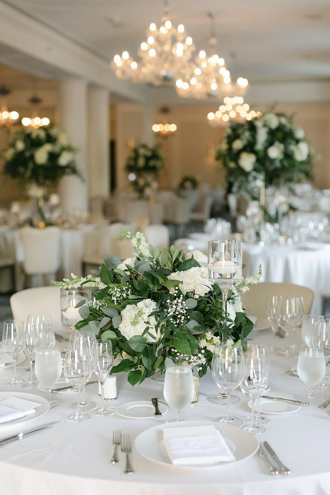 Elegant banquet table with a large floral centerpiece, surrounded by glasses, plates, and silverware in a decorated hall with chandeliers.