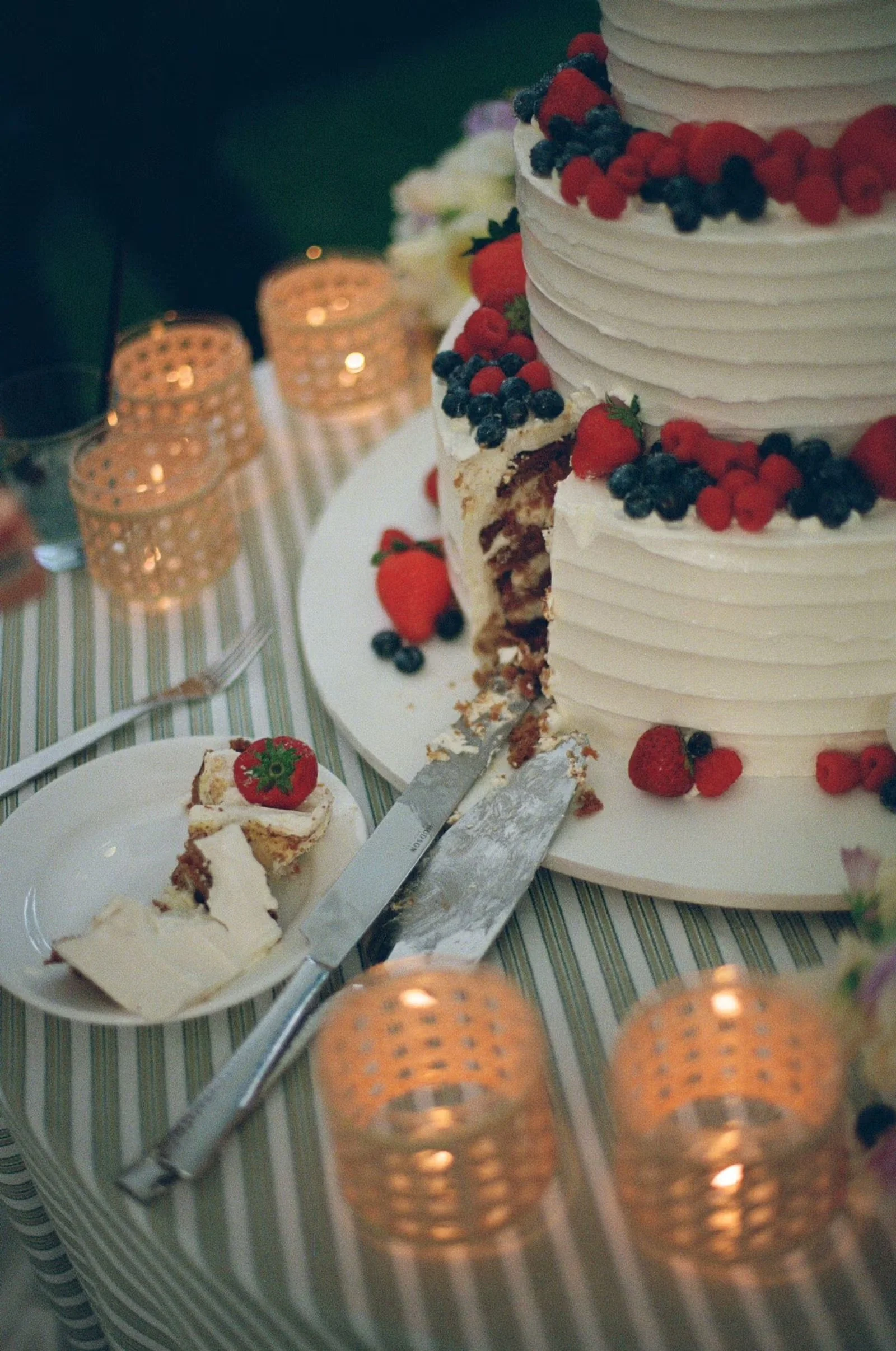 White wedding cake with strawberries, blueberries, and rassberries surrounded by warm candels on a table with a stripped tablecloth.