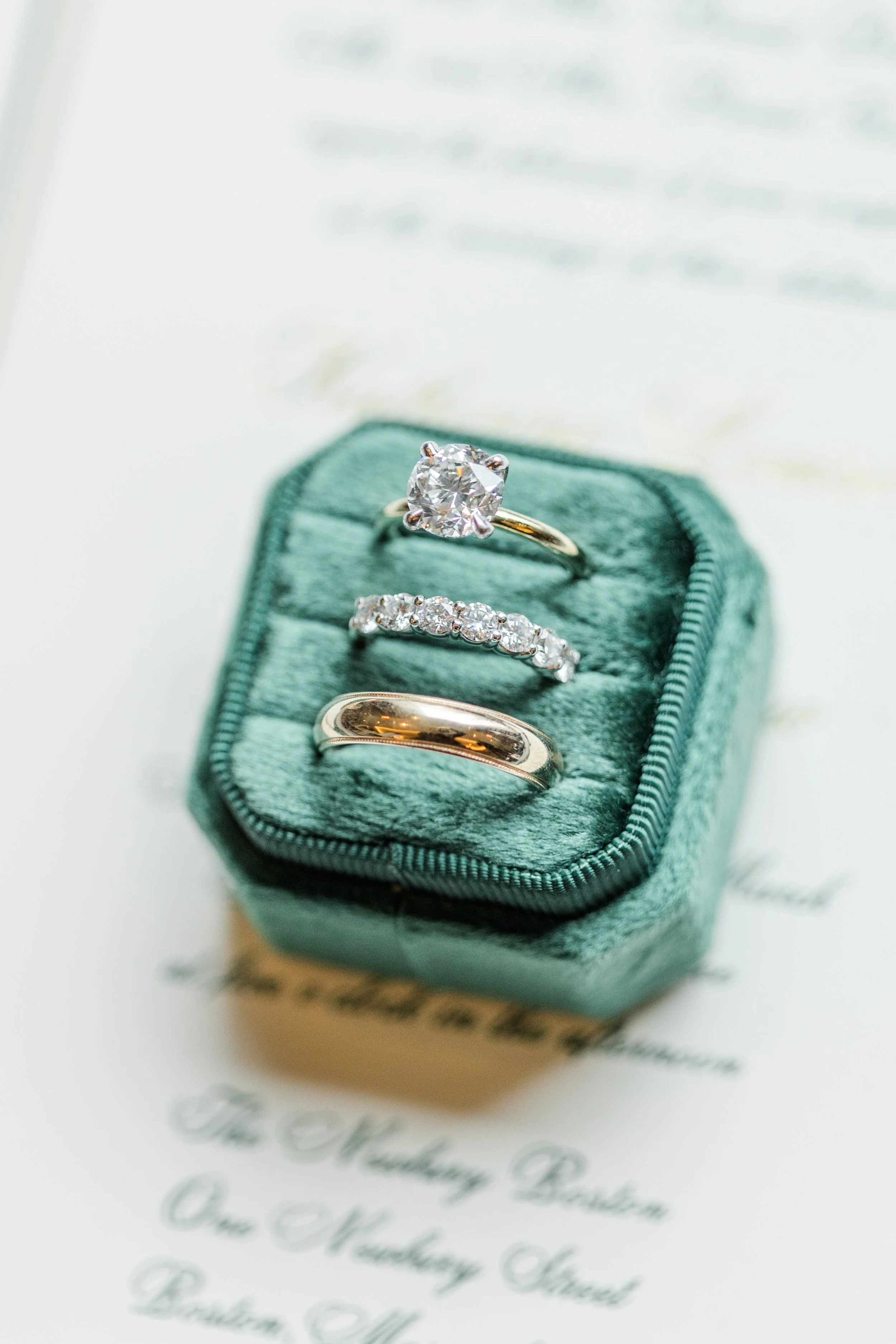 a diamond ring with a gold band, a band of diamonds, and a gold wedding band sit in a velvet emerald green ring box.
